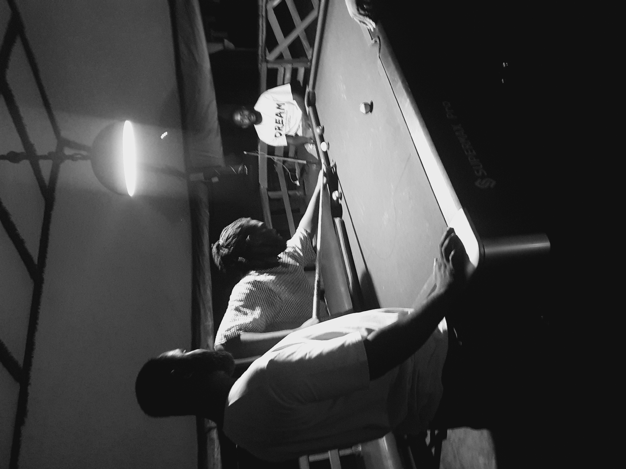 In Lokoja, Nigeria, three men are engaged in a game of billiards outdoors, under a lit lamp. The setting appears to be a dimly lit open-air space, possibly a patio or a covered area with a slatted roof. A billiard table, branded "SUPERMAX Pro," is centrally positioned. One man, wearing a white t-shirt, is in the foreground, partially silhouetted, leaning over the table and appearing to be in the process of taking a shot. Another man, dressed in a patterned shirt, is also leaning over the table, holding a cue stick, suggesting he may have just taken a shot or is preparing to. A third man is seated in the background, wearing a white t-shirt with the word "DREAM" printed on it. He is observing the game. The overall atmosphere is casual and social. The lighting is dim, with the overhead lamp casting a bright pool of light on the table and the surrounding area, creating strong shadows. The background shows wooden railings and structures, indicating an outdoor environment.
