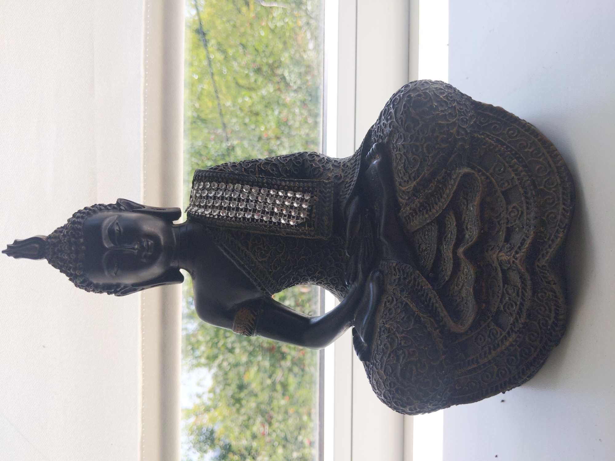 The image shows a dark, ornate statue of Buddha in a seated meditation pose, placed on a white surface in front of a window. The statue is detailed with intricate patterns on its robes and base, and a jeweled band adorns its left shoulder. Through the window, out-of-focus green foliage suggests an outdoor environment. The overall lighting is bright, coming from the window. The scene does not provide any information or visual cues that could link it to the city of Kyiv, Ukraine, or any specific urban location.