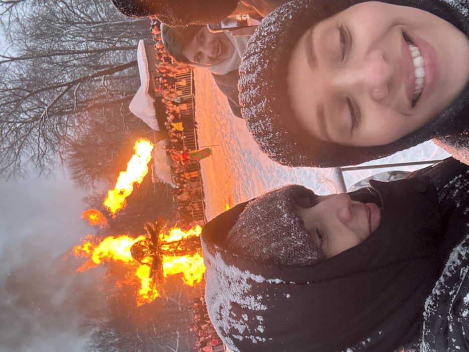 A selfie captures three people in snowy winter attire. A woman in a sparkly hat smiles widely, while another person below her wears a dark hooded jacket. A third person peeks through in the background. Behind them, a large bonfire blazes, with flames licking the sky. A crowd of onlookers can be seen gathered near the fire, along with a small white structure. The trees are bare, and snow covers the ground, adding to the winter setting.