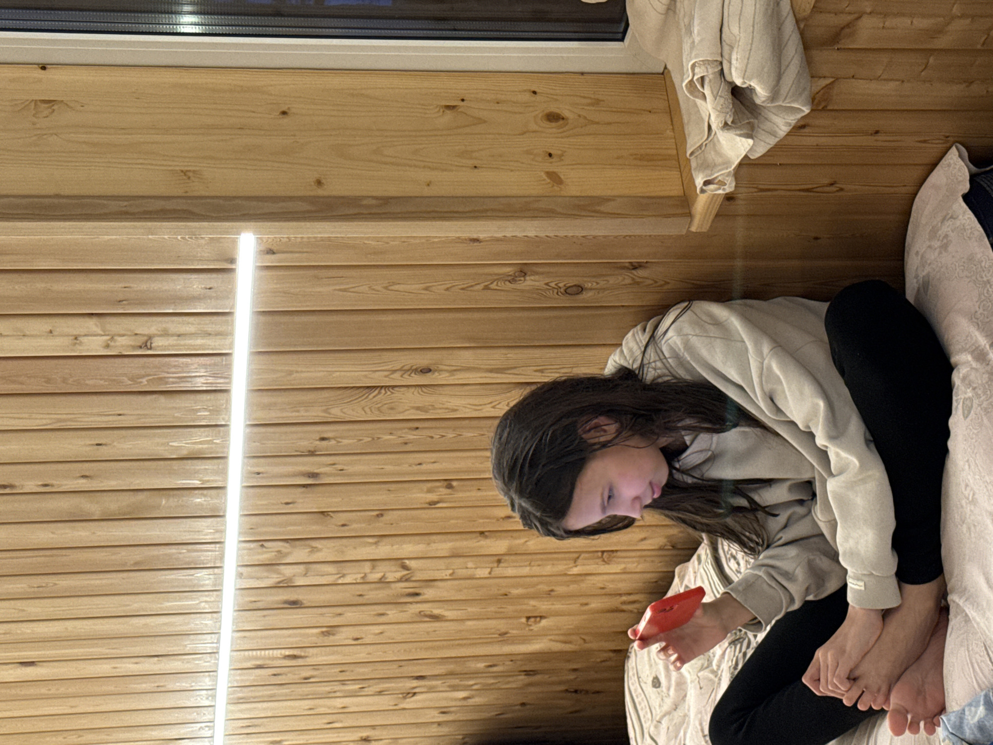 A person is seated on a bed, looking at a red device in their hands. They are wearing black pants and a light-colored hoodie. The background consists of wooden paneling, with a window at the top. A light fixture is also visible on the wall. The scene is located inside a room. There is no discernible information available within the image to indicate that this scene is in the city of Chaikovsky, Russia.