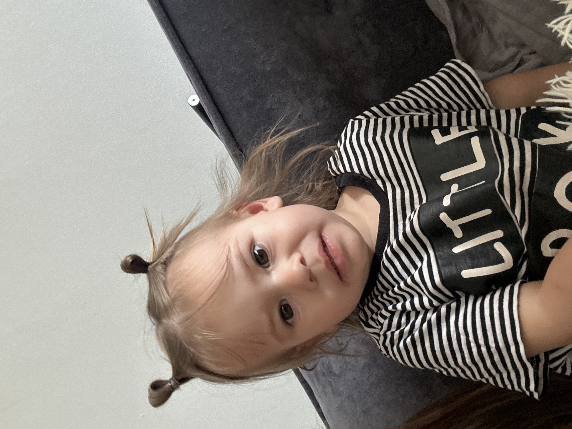 A young child lies on a dark gray upholstered surface, possibly a couch or bed, against a white wall. The child is fair-skinned with light brown hair styled in two small ponytails. She wears a black and white striped shirt with large white lettering partially visible. The environment seems domestic. The location is not evident from the image.