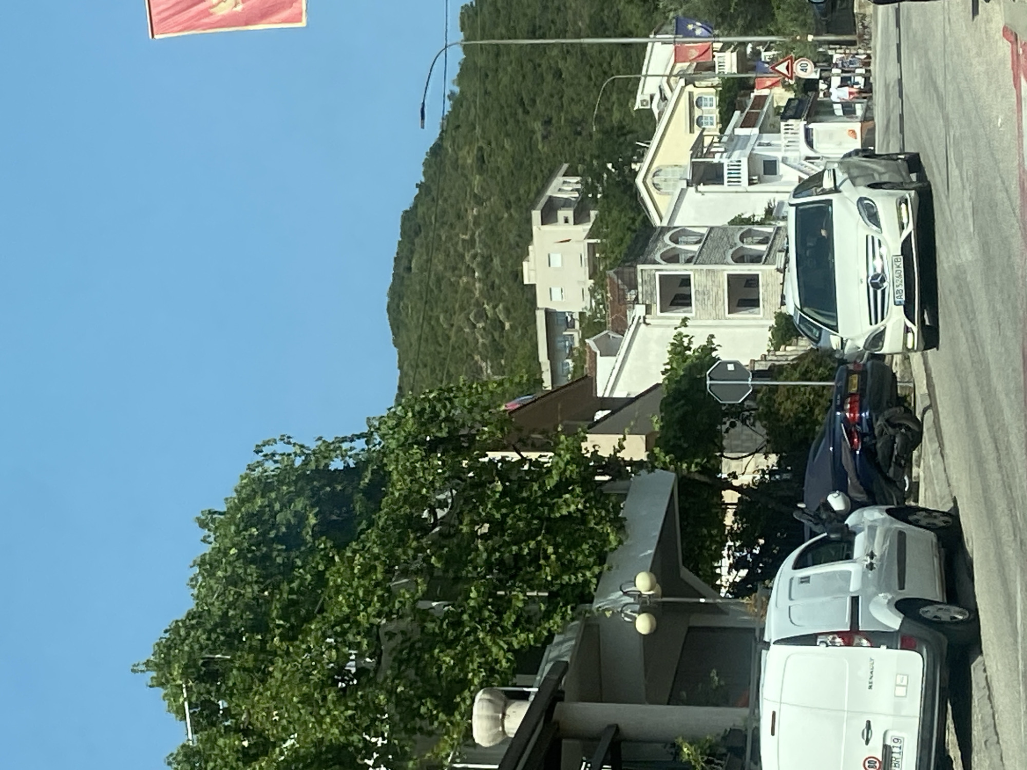 The image shows a street scene in Budva, Montenegro, on a sunny day. In the foreground, a white Renault van is parked on the left side of the road. To its right, a dark-colored car is parked, with a scooter behind it. Further down the street, a white Mercedes-Benz is driving towards the camera. Several multi-story buildings line the street, with balconies and vegetation. In the background, a lush green hill rises, dotted with more buildings. A red flag with a yellow emblem is visible at the top right of the image, possibly a Montenegrin flag. Traffic signs indicating a speed limit of 40 are also visible.