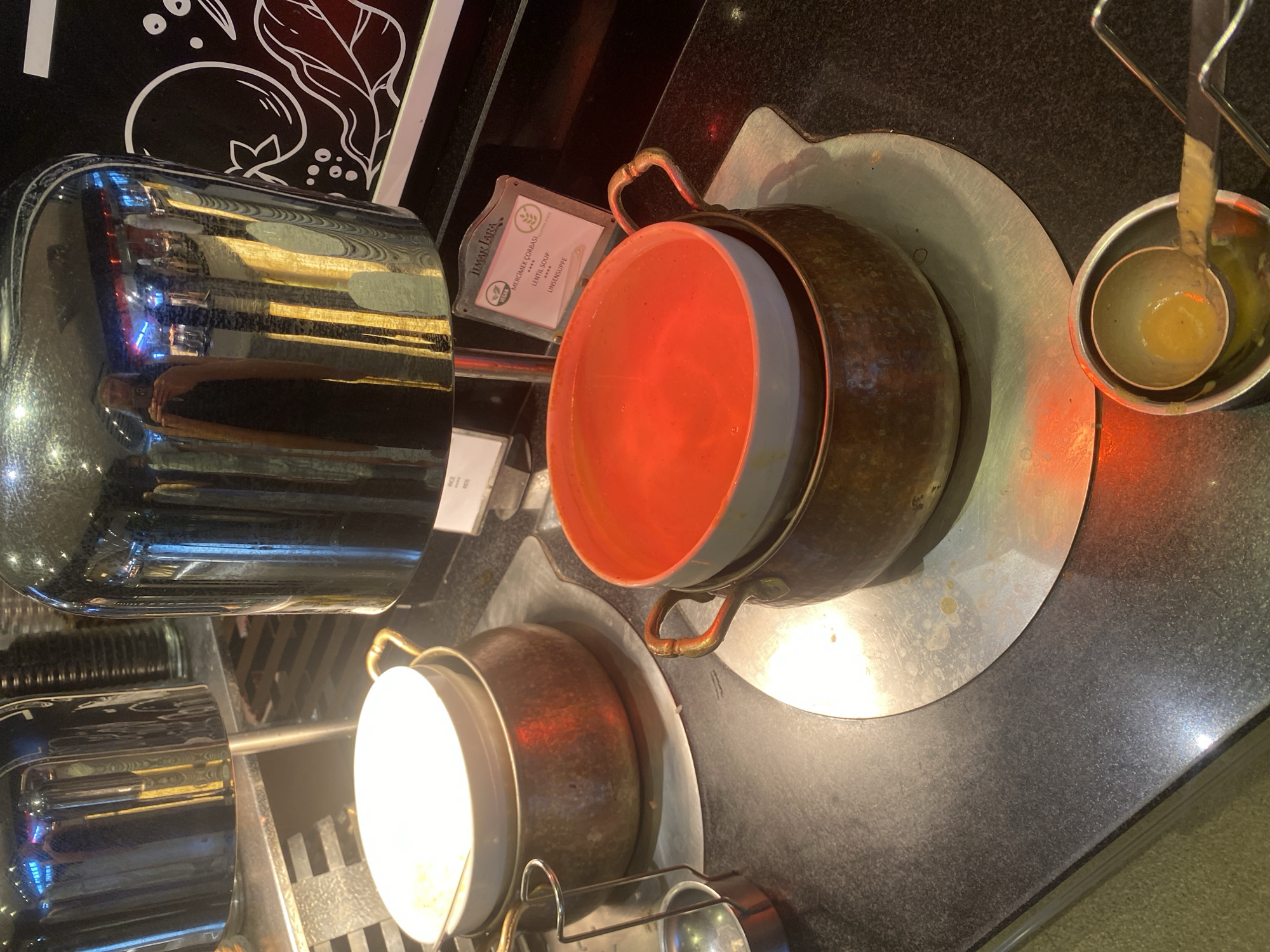 A close-up, eye-level shot shows a buffet-style food service setup with two tureens of soup. The tureen in the foreground contains a bright orange soup and is situated on a metal stand. A ladle rests in a small metal container next to the tureen. The second tureen, to the left and slightly behind, holds a lighter colored soup. Both tureens are placed on circular metal plates on a dark countertop. Above the tureens, large, reflective metallic warming domes are visible, one of which shows a reflection of a person holding a phone. A small sign in front of the orange soup tureen reads "JIMAR LARA VEGAN MERCIMEK CORBASI LENTIL SOUP LINSEN SUPPE ****". The overall environment suggests an indoor restaurant or catering facility, possibly in Aksu, Turkey, given the text on the sign. The lighting is warm and artificial.