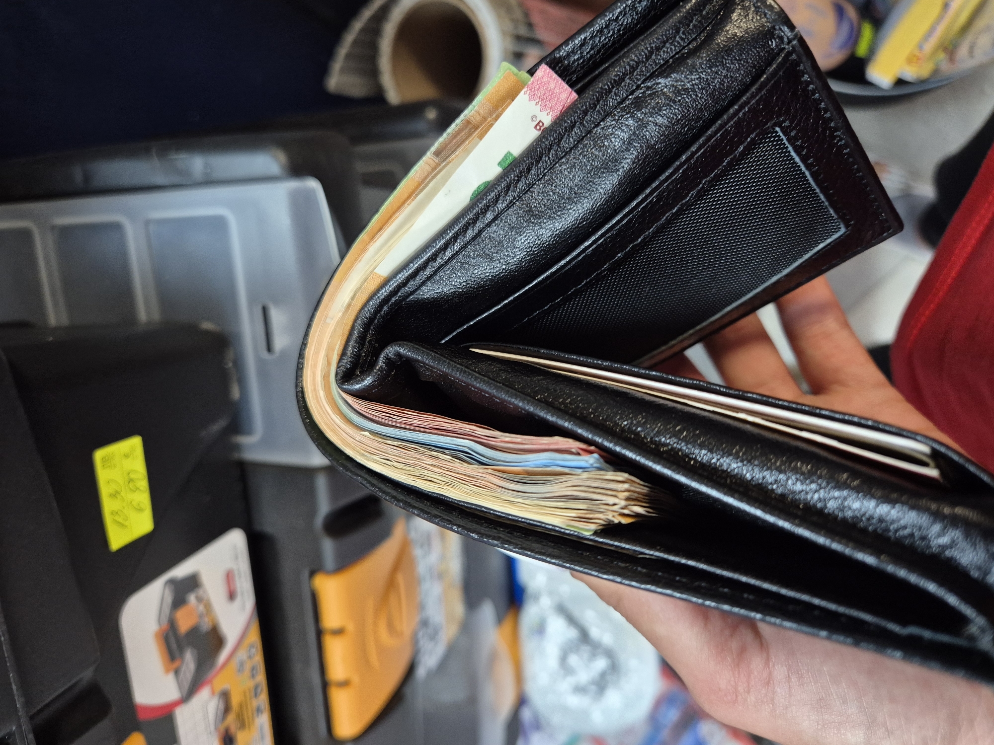 A light-skinned hand holds an open black leather bi-fold wallet in a slight upward angle. The wallet is significantly expanded, filled with a dense stack of various colored banknotes, predominantly yellow, orange, blue, and red hues, indicating multiple denominations or currencies. A portion of a green banknote with a faint 'B' or 'R' and a pink stripe is visible on the right side of the wallet.
The background, likely within a storage or work area in Iskrets, Bulgaria, features a black plastic or metal storage unit or shelving. Affixed to this unit is a yellow rectangular sticker with handwritten black text: "13.50" on the top line and "6.90 B" on the bottom line, possibly indicating prices in Bulgarian Lev. Below and to the left of the sticker, a white label with a graphic of a black and orange toolbox is partially visible. An orange-colored object is present in the mid-ground. The right side of the background shows a dark blue/grey surface and a beige-colored roll of material, possibly tape or fabric, with a corrugated texture. A red garment is partially visible at the bottom right corner of the frame. Other blurry, indistinguishable objects are present in the deeper background.