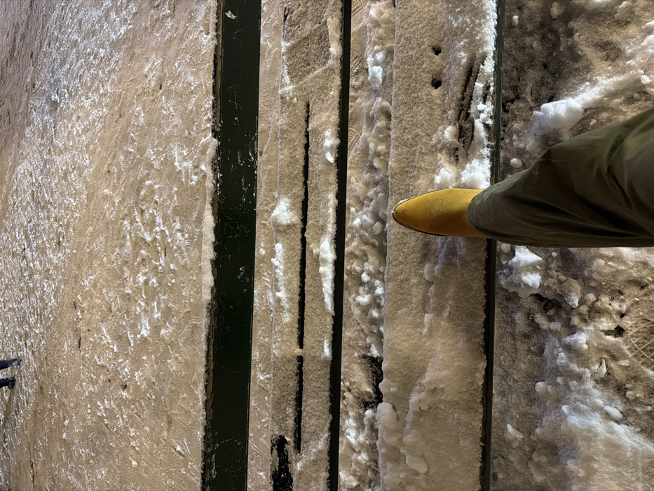 A person's leg and foot with a mustard yellow shoe rest on snow-covered steps. The steps are constructed with narrow, dark vertical supports and wider treads filled with snow. The snow exhibits varying textures, with some areas appearing fluffy and others more compacted. A portion of the bottom of a railing is visible on the left. The overall scene suggests a winter setting with the person potentially navigating a snowy staircase.