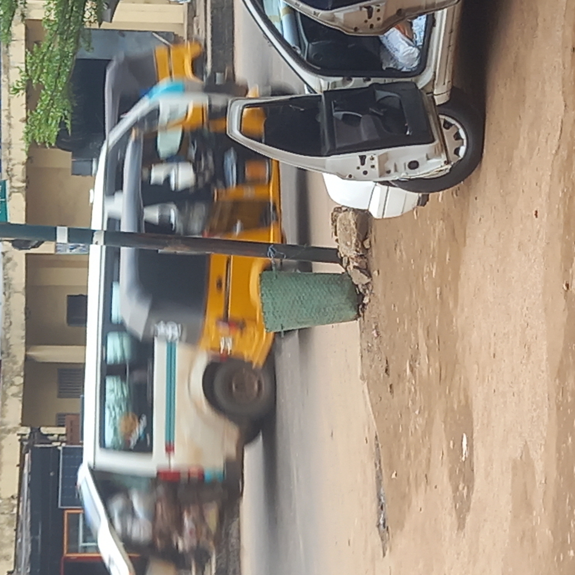 A three-wheeled vehicle, with its door open, is parked on a dirt and concrete road in Dadi Riba, Nigeria. Another similar vehicle, yellow and white, moves along the road in the background. A green, cylindrical object is attached to a post. The scene includes portions of nearby buildings.