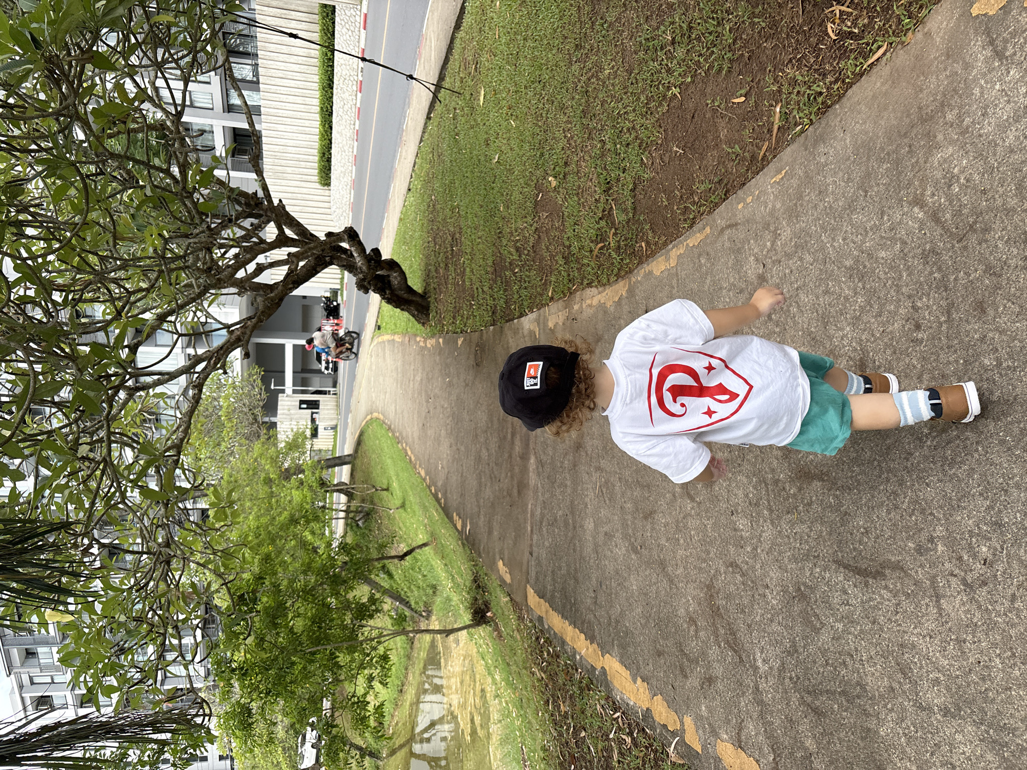In the city of Choeng Thale, Thailand, a young child, viewed from behind, walks along a concrete pathway lined with yellow paint markings. The child wears a black baseball cap, a white t-shirt with a red emblem on the back, teal shorts, and striped socks with brown shoes. To the left of the pathway is a body of water with a greenish tint and grassy banks dotted with young trees. To the right of the pathway is a strip of grass and then a road with a white concrete barrier and red and white painted curbs. A motorbike with two people riding on it is visible on the road in the distance. Behind the road, there is a multi-story building with modern architecture. Overhead, the branches and leaves of a large tree partially obscure the sky.