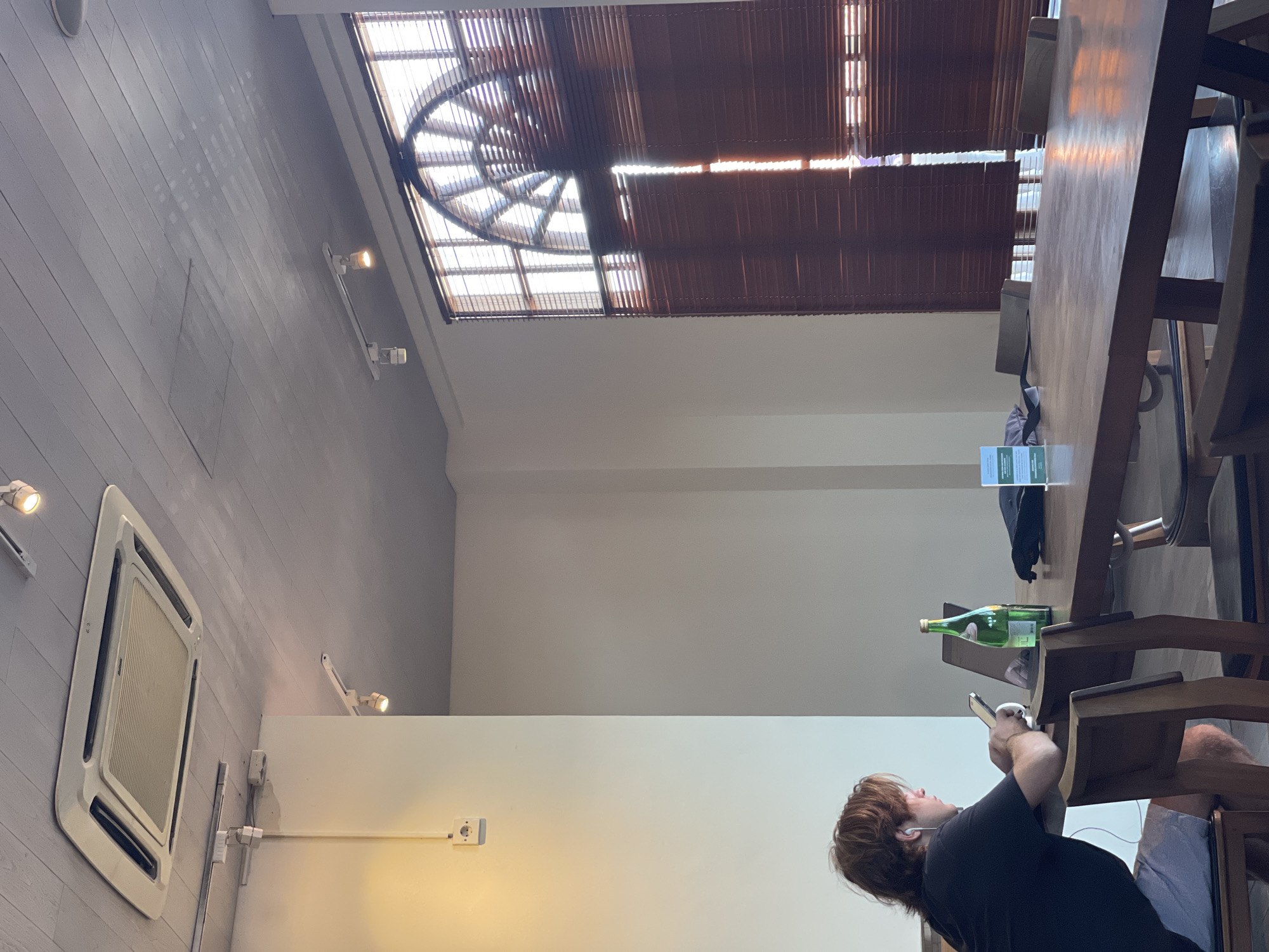 A person is looking at their phone while seated at a wooden table in what appears to be a cafe or restaurant. On the table are a green bottle and some papers. The cafe has a unique ceiling structure with a combination of open sections and dark wooden beams. There is an air conditioning unit on the wall. The location is North Kuta, Indonesia.