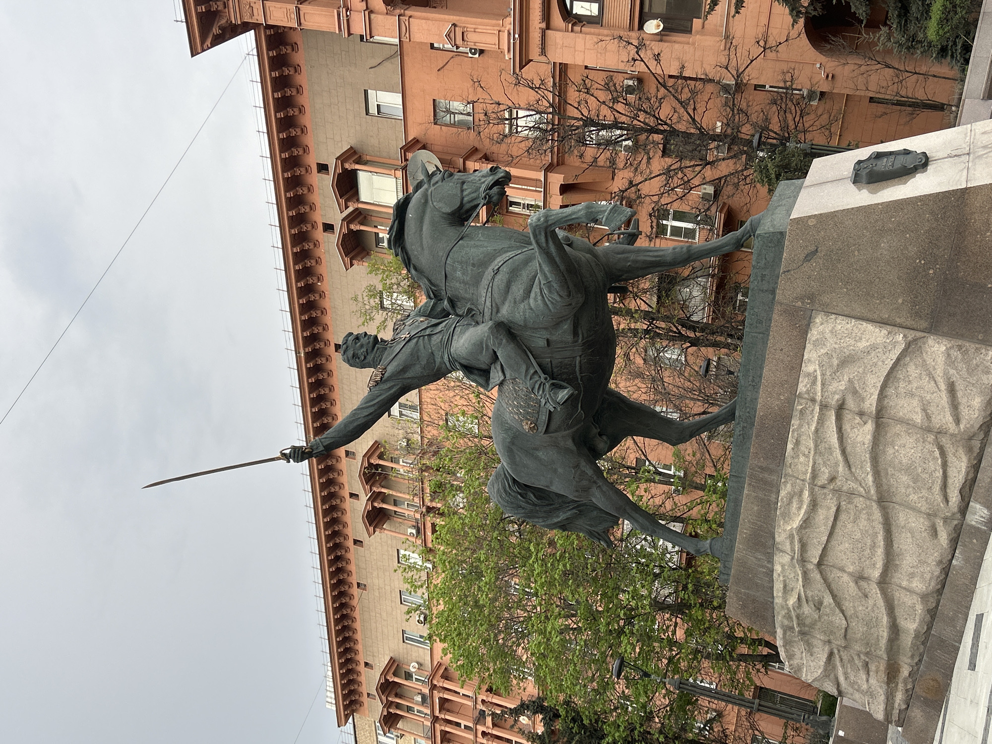 Here is a description of the image:
The image shows a statue of a man on horseback, holding a sword. The statue is dark and appears to be made of metal. The horse is rearing up, and the man is pointing the sword forward. In the background, there is a large building with many windows. There are also trees with green leaves. The sky is overcast. The statue sits atop a granite base. The statue location is in the city of Unknown, Unknown.
