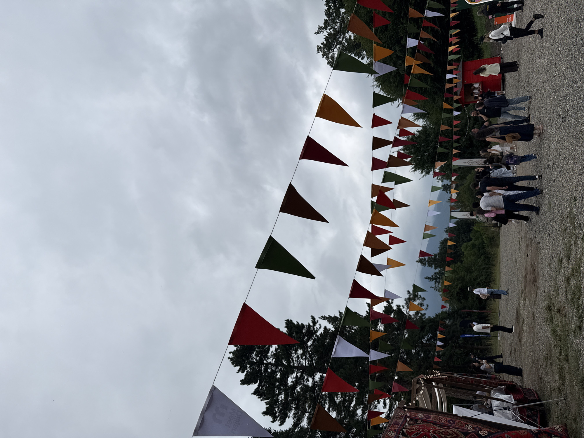 The image depicts an outdoor market or festival in Dilijan, Armenia, under a cloudy sky. Colorful triangular flags are strung across the scene, creating a festive atmosphere. Several people are visible, some walking and others gathered in small groups. In the foreground to the left, a stall with a patterned textile covering and a white canopy is set up. Further back, a small red kiosk is visible, possibly for food or beverages. The ground is covered with gravel, and a dense forest with tall trees forms the backdrop, with hills or mountains in the distance. The overall impression is of a community gathering in a natural setting.