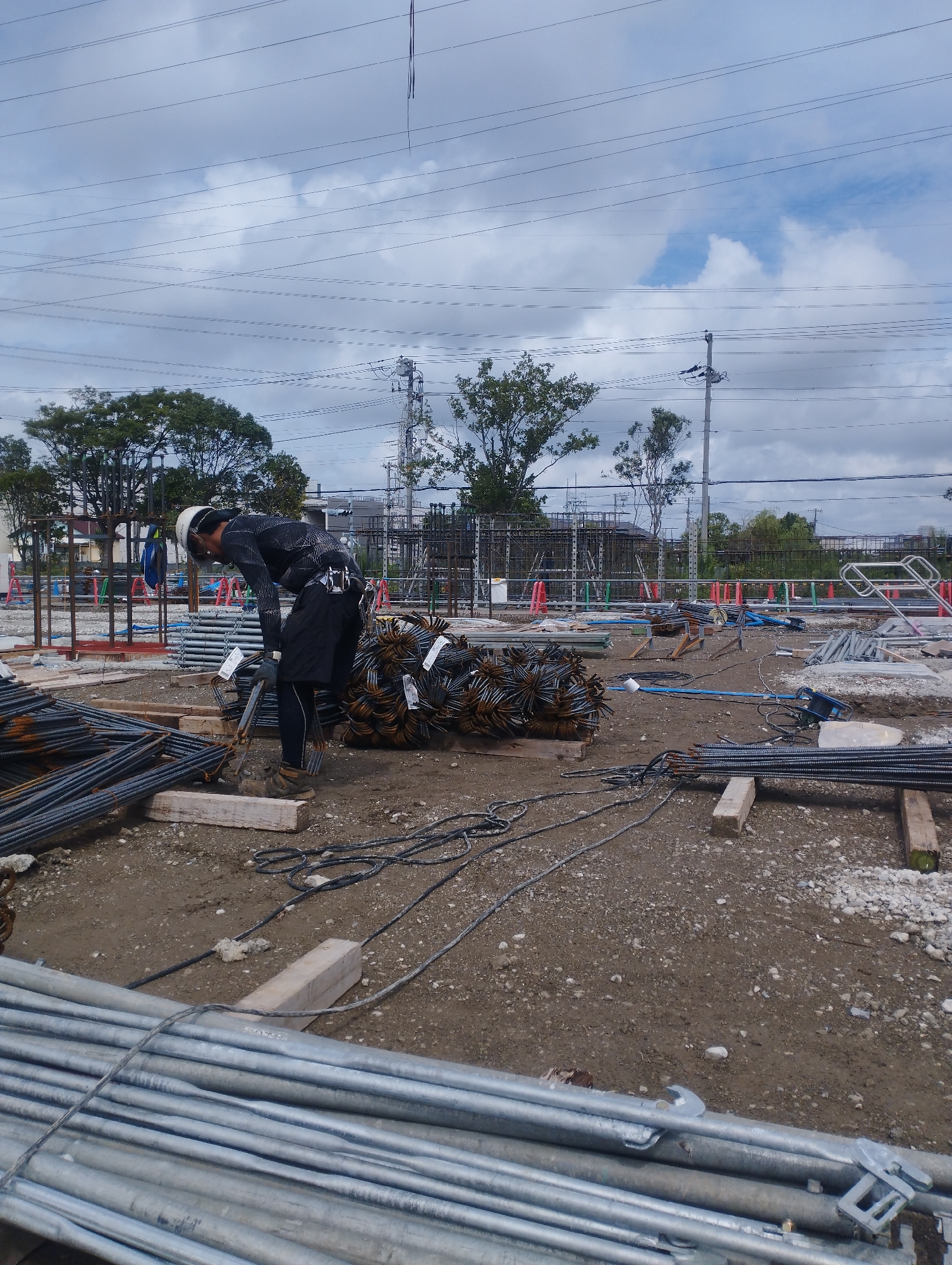 The image depicts a construction site in Ichikawa, Japan, likely a portion of the Chiba Expressway construction project. In the foreground, a male construction worker, wearing a white hard hat, a black long-sleeved shirt, black shorts, and work boots, is bent over, tending to bundles of steel rebar. He is equipped with gloves and appears to be securing or organizing the rebar. The ground is a mix of dirt and gravel, with various construction materials scattered around, including more piles of rebar, wooden beams, coiled cables, and sections of metal piping. In the midground, there are more organized stacks of steel rebar and scaffolding structures. In the background, a series of tall utility poles with numerous power lines crisscross the sky, a common sight in urban and industrial areas of Japan. Trees and buildings are visible beyond the construction zone, suggesting an urban or suburban setting. The sky is overcast with scattered clouds. The overall scene is one of active construction work in an outdoor environment.