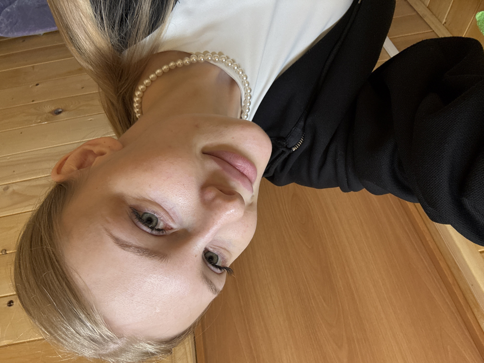 The image depicts a young adult female captured in a close-up selfie indoors in Perm, Russia. She has blonde hair, pulled back into a ponytail visible over her right shoulder, and a light complexion with visible pores. Her green eyes are directed towards the camera, with prominent long eyelashes. Her full lips are naturally colored and slightly parted in a mild smile. She is wearing a white crew-neck top underneath a black textured, zippered hooded jacket. A choker-style pearl necklace, composed of uniformly sized spherical pearls, encircles her neck. The background consists of light to medium brown wooden paneling, arranged both horizontally and vertically, with a visible wood grain pattern. A portion of her left arm, covered by the black jacket, is visible in the lower left corner, indicating she is holding the camera.