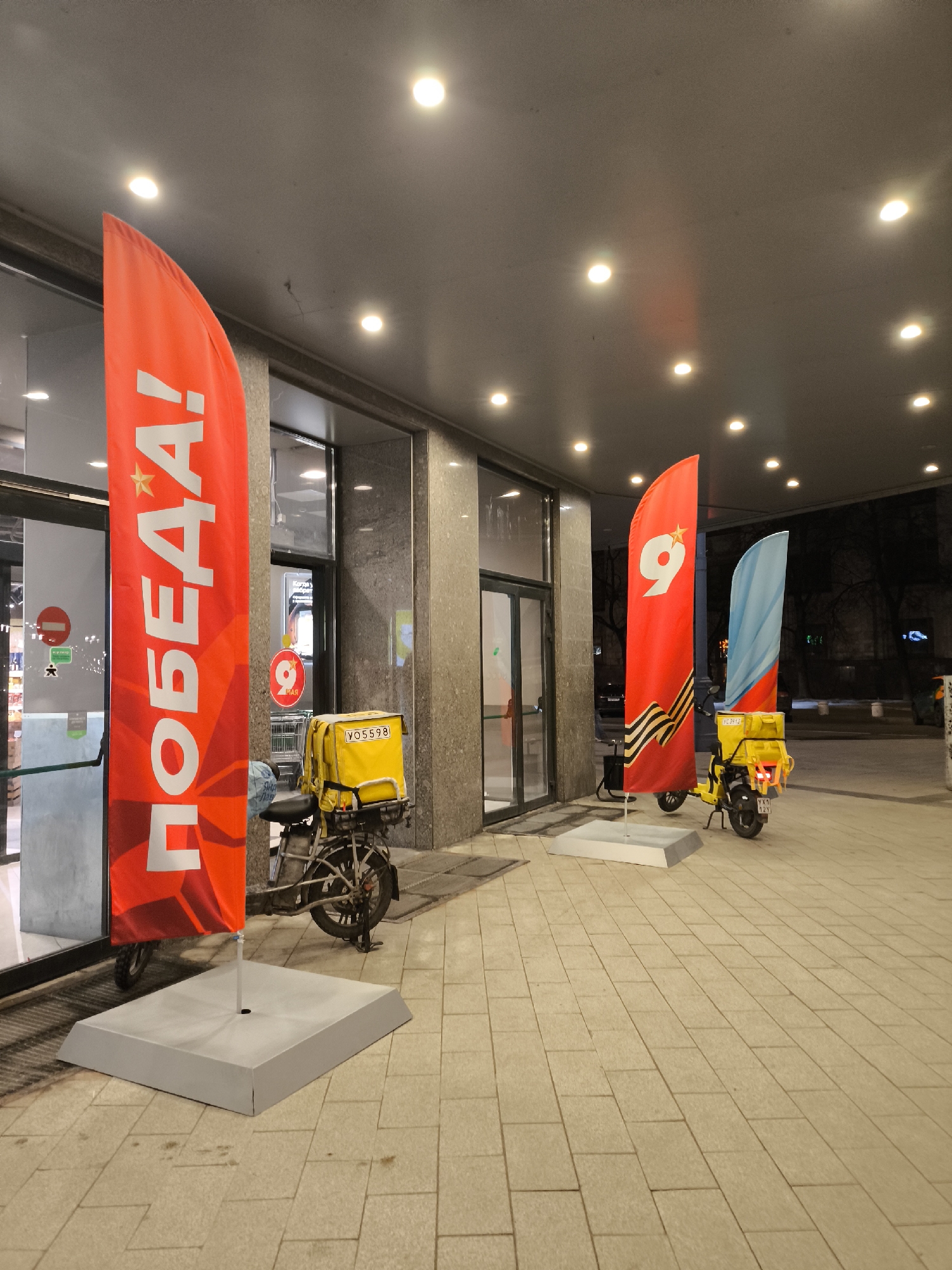 The image displays an outdoor scene at night in front of a modern building in Moscow, Russia. The area is illuminated by numerous recessed lights embedded in the ceiling of an overhead canopy. The ground is paved with light-colored, rectangular tiles.
The main subjects are three large, vertical flags and two delivery mopeds, positioned on the paved area near the building's entrance. The building itself features a dark grey, possibly granite, facade with large glass entrance doors.
On the far left, a red flag prominently displays white Cyrillic text "ПОБЕДА!" (Victory!) with a star above it. The middle flag is also red, featuring a large white number "9" with a star, and a depiction of a St. George's ribbon at its base. The flag on the right is light blue with a red stripe and an emblem, though less distinct.
Two yellow delivery boxes, characteristic of food or package delivery services, are mounted on electric mopeds. One moped is situated near the left flag, its yellow box bearing the identifier "УО 5598". Another moped is positioned further right, between the middle and right flags, with its license plate "УХ 12У" visible.
Through the glass doors of the building, a circular red sign with a white "9" is visible, suggesting a commercial establishment that might be celebrating or commemorating an event related to the number 9, consistent with Victory Day (May 9th) celebrations in Russia. No people are visible in the scene. The time of day is clearly night due to the reliance on artificial lighting and the dark background beyond the building.