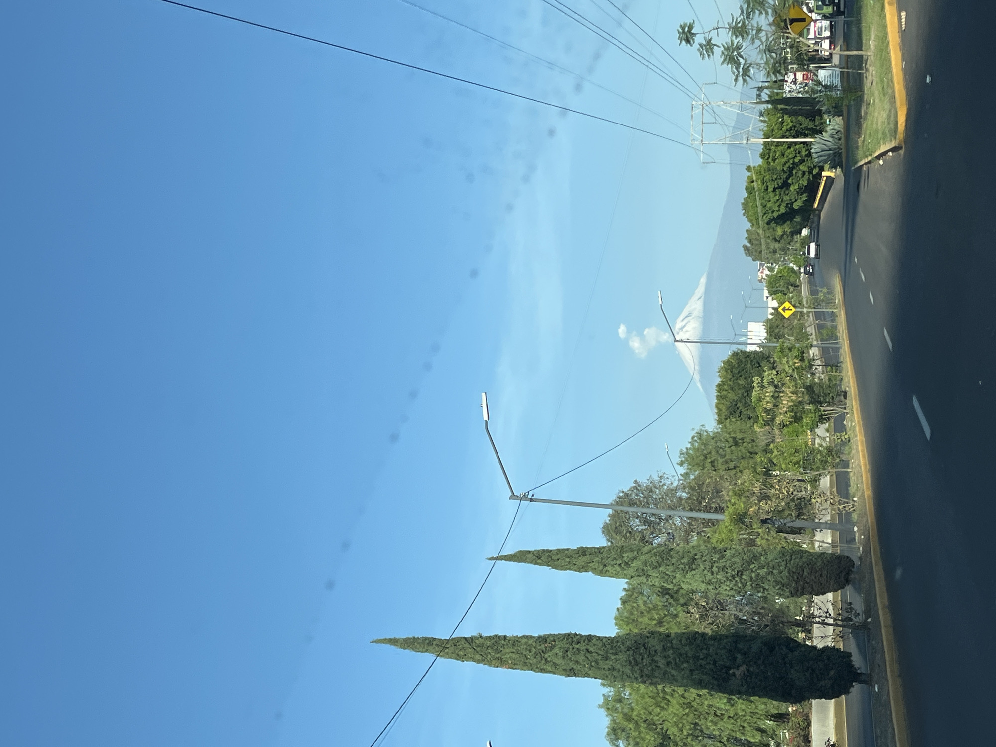 The image depicts a road in Puebla, Mexico, with a view of the Popocatépetl volcano in the distance. The sky is clear blue. Power lines stretch across the upper portion of the frame. The right side of the image shows the asphalt road surface and yellow shoulder line. Trees and streetlights are visible along the roadside. In the distance, the volcano's snow-capped peak is visible, with a plume of smoke or steam rising from its crater. Other vehicles appear on the road further away.