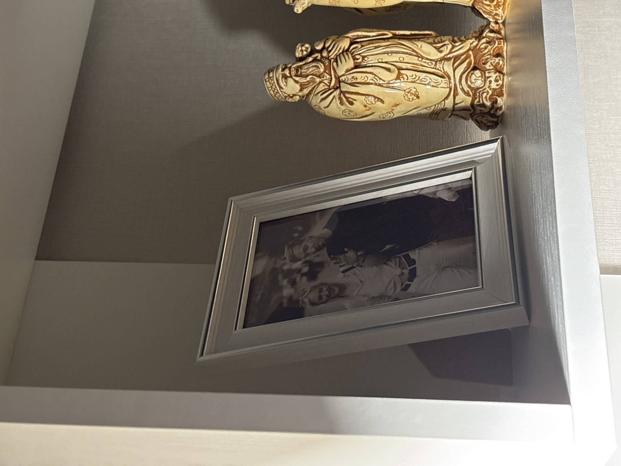 A framed black and white photograph of two people is displayed on a shelf. To the right of the photograph, there are two decorative statues. The statue on the left is of an elderly man with a beard, wearing ornate robes and holding a child. The statue on the right is partially visible. The shelf is white and appears to be part of a larger unit, possibly a bookcase or display cabinet. The background is a textured grey wall. The lighting suggests an indoor setting, likely a home. No specific location in Sarov, Russia can be identified from the image.