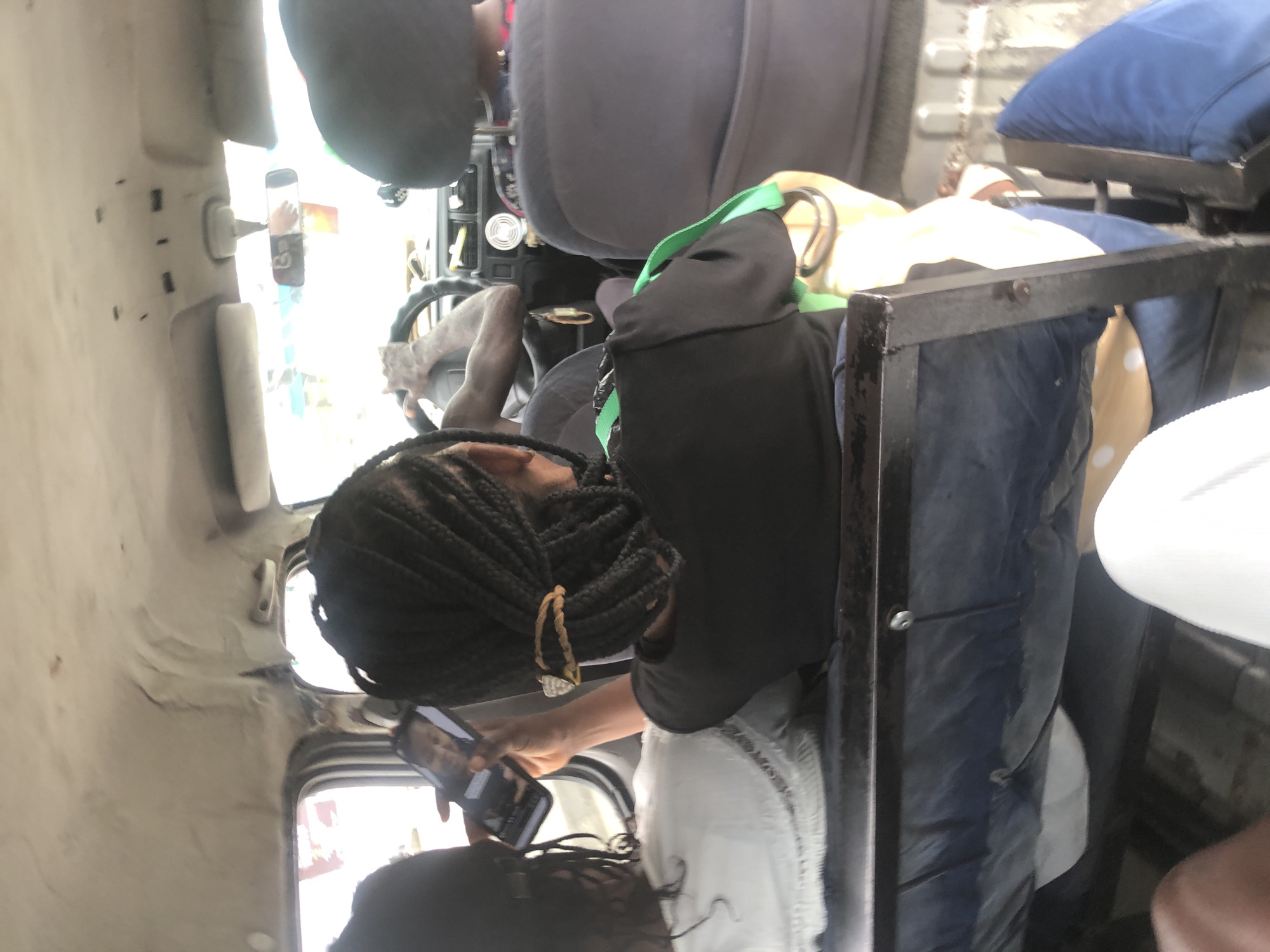 The image shows the interior of a van, likely a passenger transport vehicle, in Ado, Nigeria. The perspective is from inside the van, looking towards the front. In the foreground, a woman with braided hair is visible from the back, holding a smartphone and looking at it. She is wearing a black shirt and a yellow and green bag is beside her. To her left, another person, also seen from the back, has their hair in braids and is wearing a white garment. Further ahead, the driver of the van is visible, with their hands on the steering wheel. The van's interior appears worn, with a stained ceiling and the seats are covered in dark fabric. Outside the van, through the windows, a blurry, bright environment suggests it is daytime. The presence of multiple passengers and the van's design indicate it is being used for public transportation.