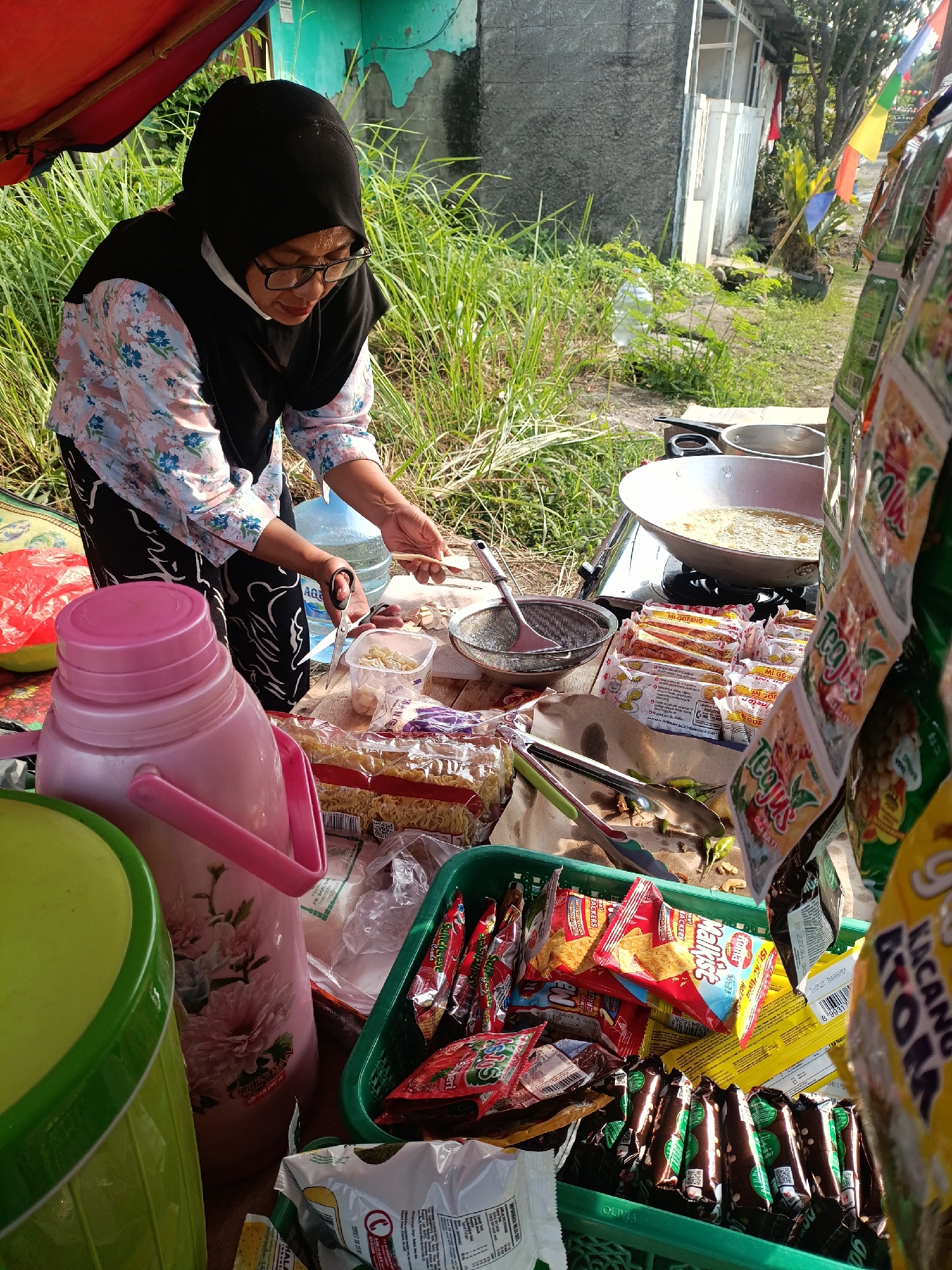 In Cariu, Indonesia, a woman wearing a black hijab and glasses, a floral patterned shirt, and a black and white patterned skirt stands at a stall. She is preparing food, using a small pair of scissors to cut something and holding a spoon. In front of her, a large pink thermos with a floral design sits next to a green bucket. Behind her, a water dispenser is visible. The stall is laden with various food items, including packets of instant noodles, snacks, and packaged seasonings. A large wok filled with frying oil is on a stove. The scene is set outdoors, with tall grass and trees in the background, suggesting a rural or semi-urban environment. The overall impression is of a small, bustling food stall in a local Indonesian setting.