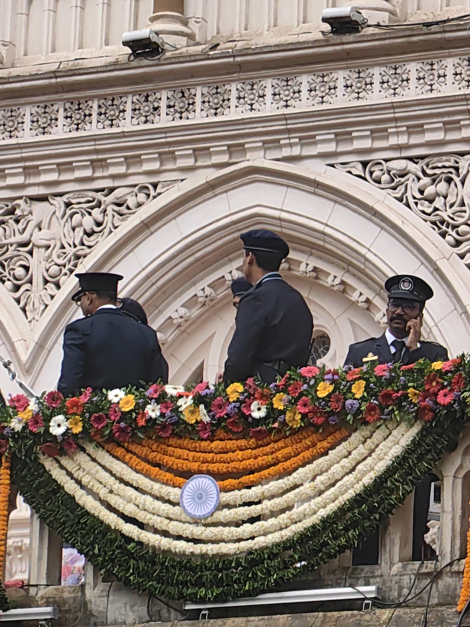 The image depicts three men in dark uniforms and caps standing on a balcony adorned with garlands of flowers. The balcony overlooks a decorative stone facade with arched openings. The man on the right is wearing glasses and appears to be speaking on a mobile phone. The floral decorations consist of a dense arrangement of colorful flowers at the top, followed by three layered garlands in white, orange, and green, with a circular emblem resembling the Ashoka Chakra in white on the middle white garland. The architectural style suggests a historical building, possibly in a public space. The scene is likely at the Chhatrapati Shivaji Maharaj Terminus (CSMT) in Mumbai, India, given its distinctive architecture and the prevalence of such floral decorations during national events.