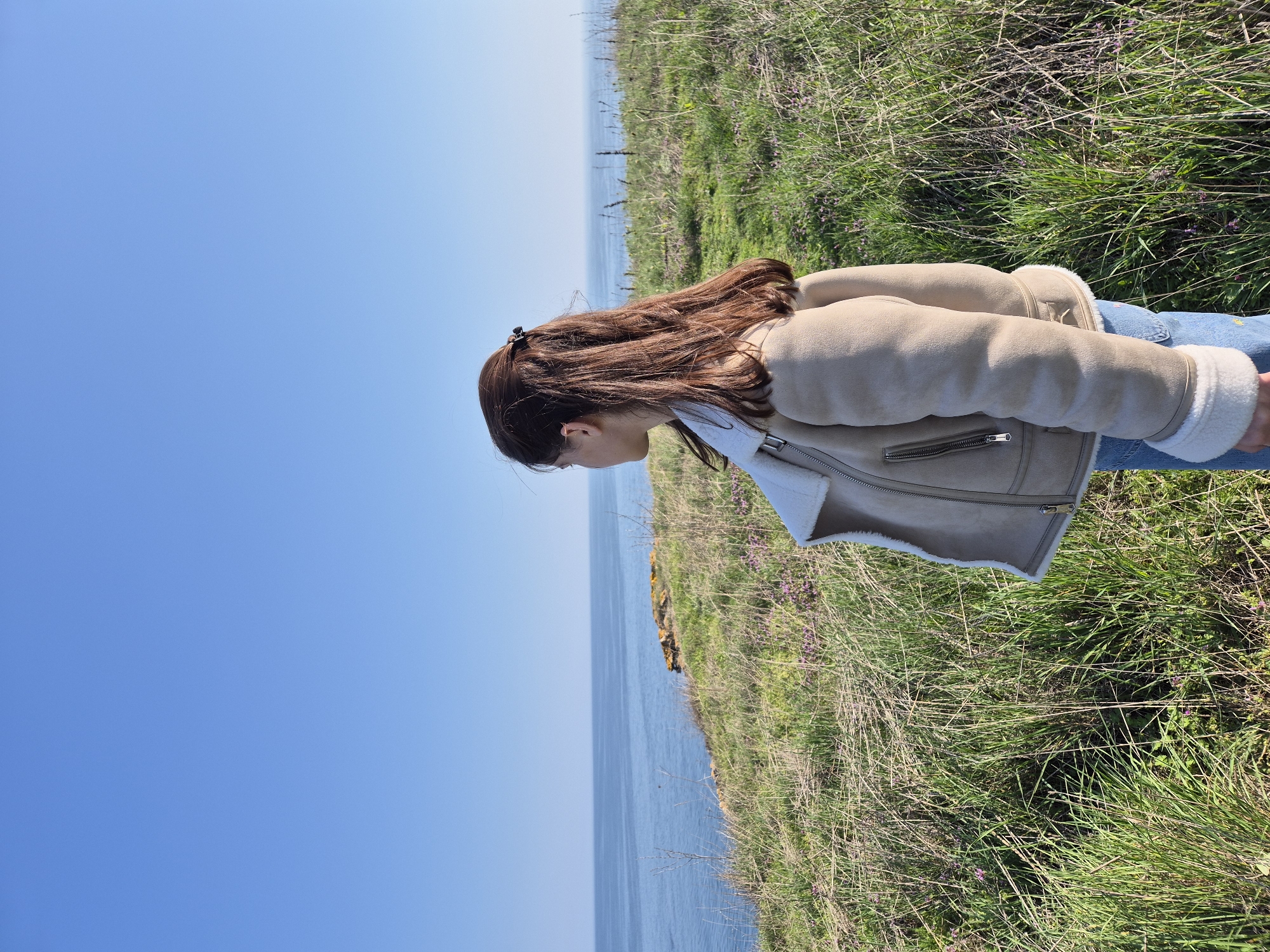 A young woman stands on a grassy cliff overlooking the Black Sea on a sunny day. The location appears to be Sveti Nikola, Bulgaria. The woman is wearing a beige jacket with white fur lining and blue jeans. She has brown hair and is looking down. The grass on the cliff is green and brown, and there are some small purple flowers growing in it. The sky is blue.
