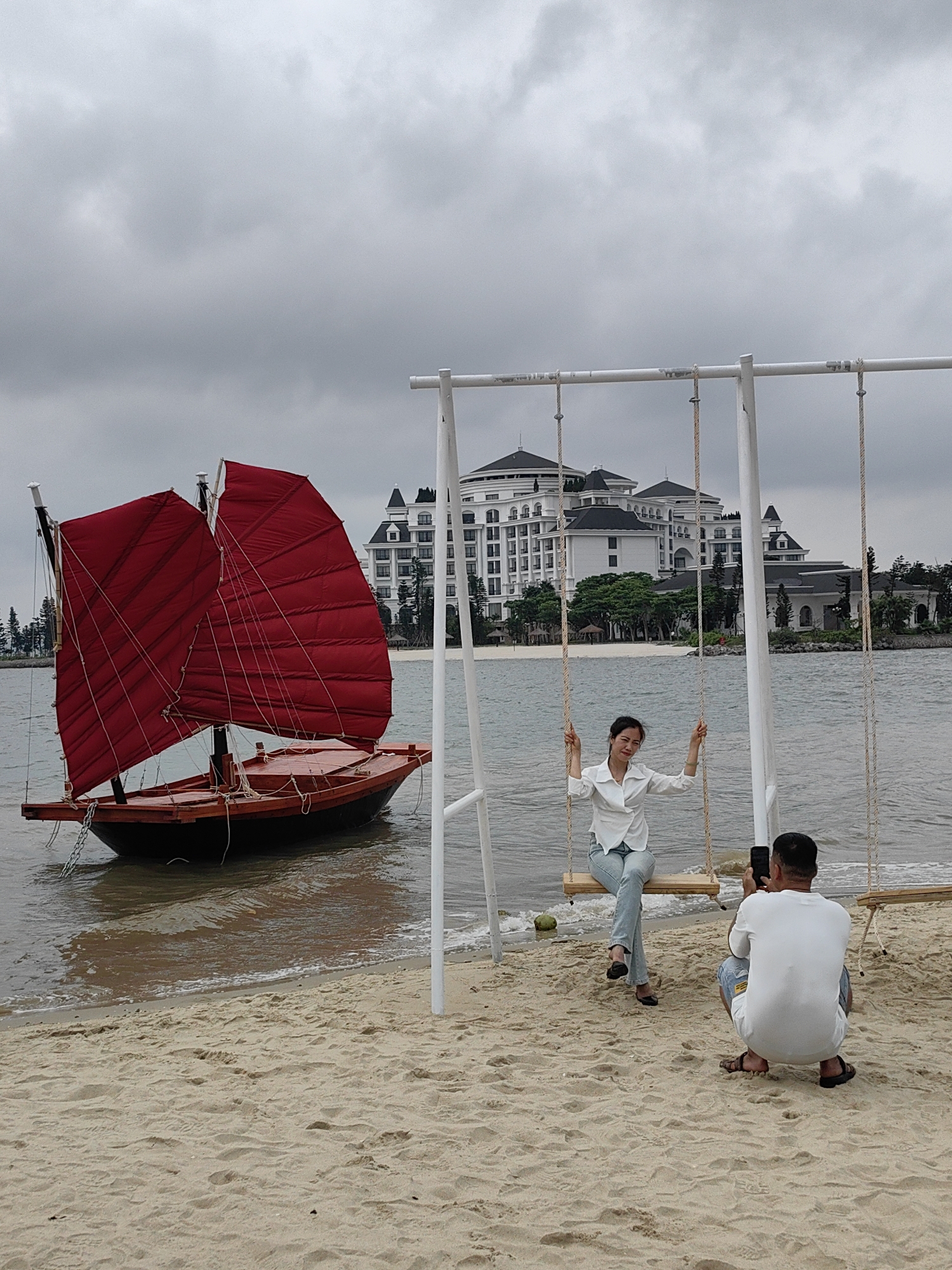 The photo was taken in Bai Chay Ward, Vietnam, at a beach. A woman in a white blouse and jeans sits on a wooden swing. A man crouches, holding a smartphone to take her picture. Behind them is a red-sailed boat in the water near the shore. Further back, a large white building is visible against a cloudy sky. The ground is sandy.