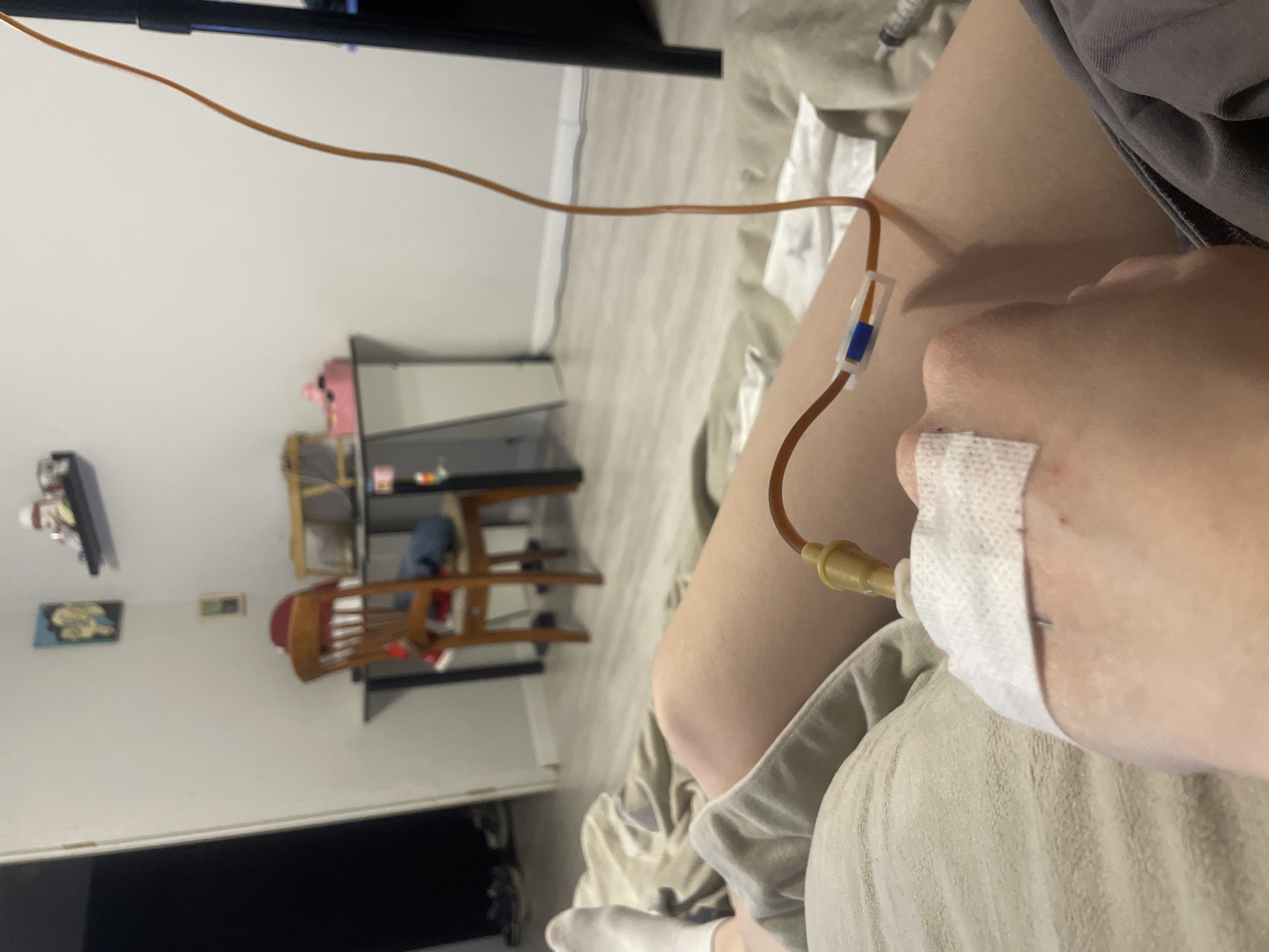 The image captures a first-person perspective of a person's body receiving an intravenous (IV) infusion. The main subject is the person's right arm, where a brown IV tube is inserted, secured with white medical tape near the wrist area. The tube runs upwards and features a blue and white flow regulator. The person's upper leg is visible on the right, covered by a gray garment, and a white sock on a foot is partially visible at the bottom.
In the background, the scene appears to be an indoor room, possibly a bedroom or living space, as indicated by various furniture items. These include a dark-colored shelf unit on the left with indistinct items, and a light-colored table with a wooden chair to the right. A pink pig figurine is visible on one of the shelves. A small framed picture with yellow details is affixed to a white wall. The room appears brightly lit, suggesting either daytime or well-lit artificial lighting. Part of a black screen or monitor is visible at the top and bottom edges of the frame. The perspective suggests the person is lying down or reclining while receiving the infusion. No visible text is present in the image.