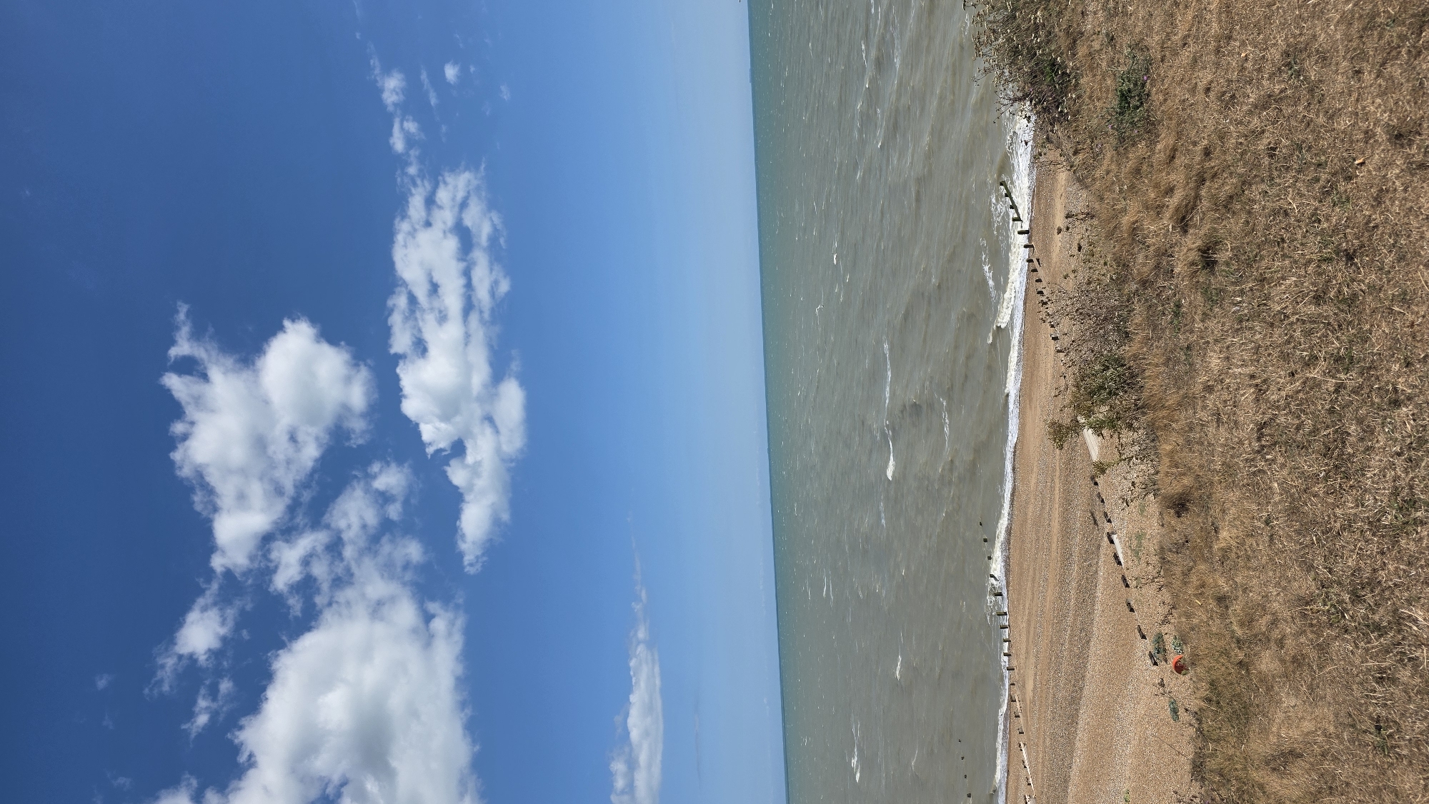 The image shows a wide shot of a sea on a clear day. The sky is bright blue with scattered white clouds. The sea is a muted green-grey color, with small waves rolling towards a pebble beach. Wooden groynes, spaced at intervals, break the waves as they approach the shore. The beach is made up of smooth pebbles and rocks, with some patches of dry grass and vegetation on the hillside overlooking it. The location is the coast of Rother, United Kingdom, a region known for its scenic coastline. There are no people or animals visible in the image. The overall impression is one of a natural, windswept coastal landscape.