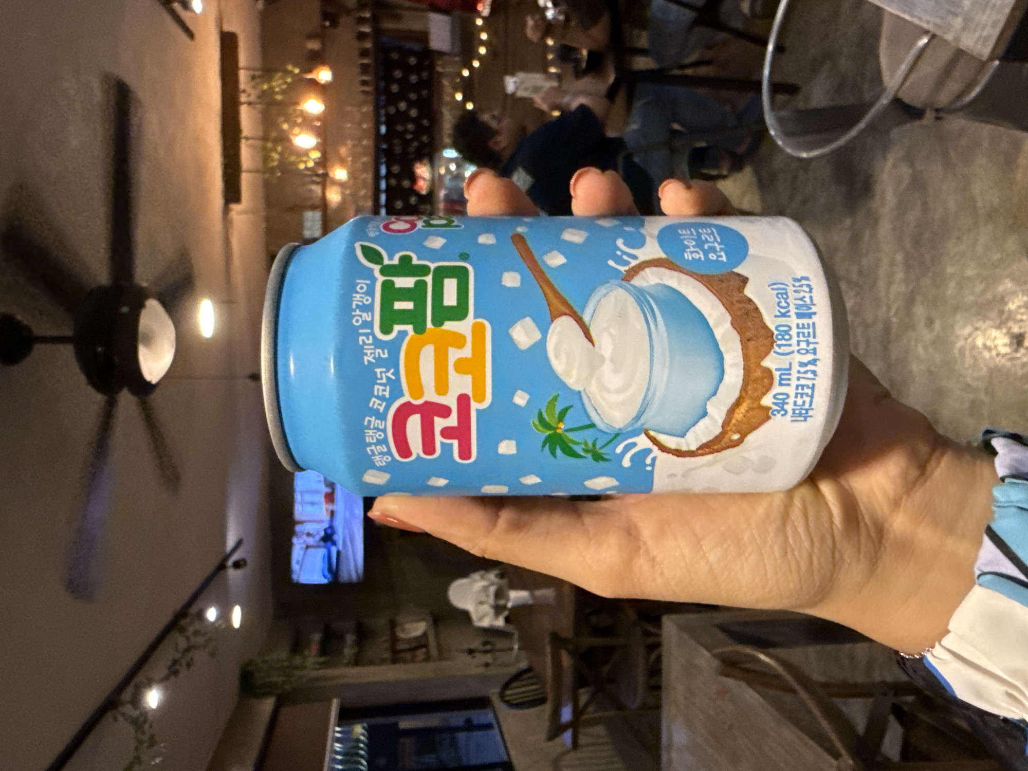 A person is holding a can of coconut drink with the brand name "Koko Palm" in Korean. The drink has an image of a coconut with a spoon in it. The person is indoors, possibly in a cafe or restaurant, as there are tables, chairs, and other people in the background. A ceiling fan is visible. Based on other information, this location is potentially in the city of Puebla, Mexico.