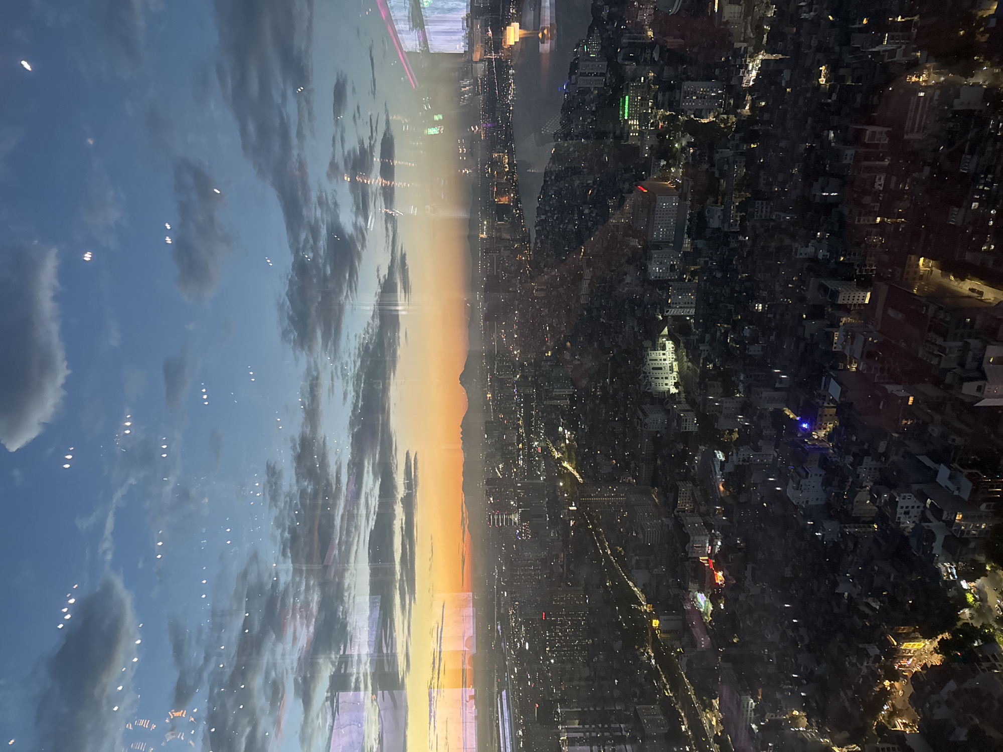 This is an aerial shot of Hanoi, Vietnam, taken during twilight. The image is vertically oriented. On the left side, a dusky sky with clouds and scattered lights reflects on a surface, possibly a window. The sky transitions from a deep blue to a warm orange glow near the horizon, with faint outlines of distant mountains visible.
The right side of the image shows a densely populated urban landscape at night. Countless lights illuminate the buildings and streets, indicating a bustling city. Various structures, from small houses to larger buildings, are visible, interspersed with patches of darkness that could be trees or unlit areas. Some buildings stand out with their illuminated facades and windows.
The scene captures the transition from day to night over a large city. The lighting suggests it's either sunrise or sunset, with the fading natural light giving way to the artificial lights of the city. The perspective is from a high vantage point, offering a sweeping view of Hanoi's urban sprawl. There is no visible text in the image.
