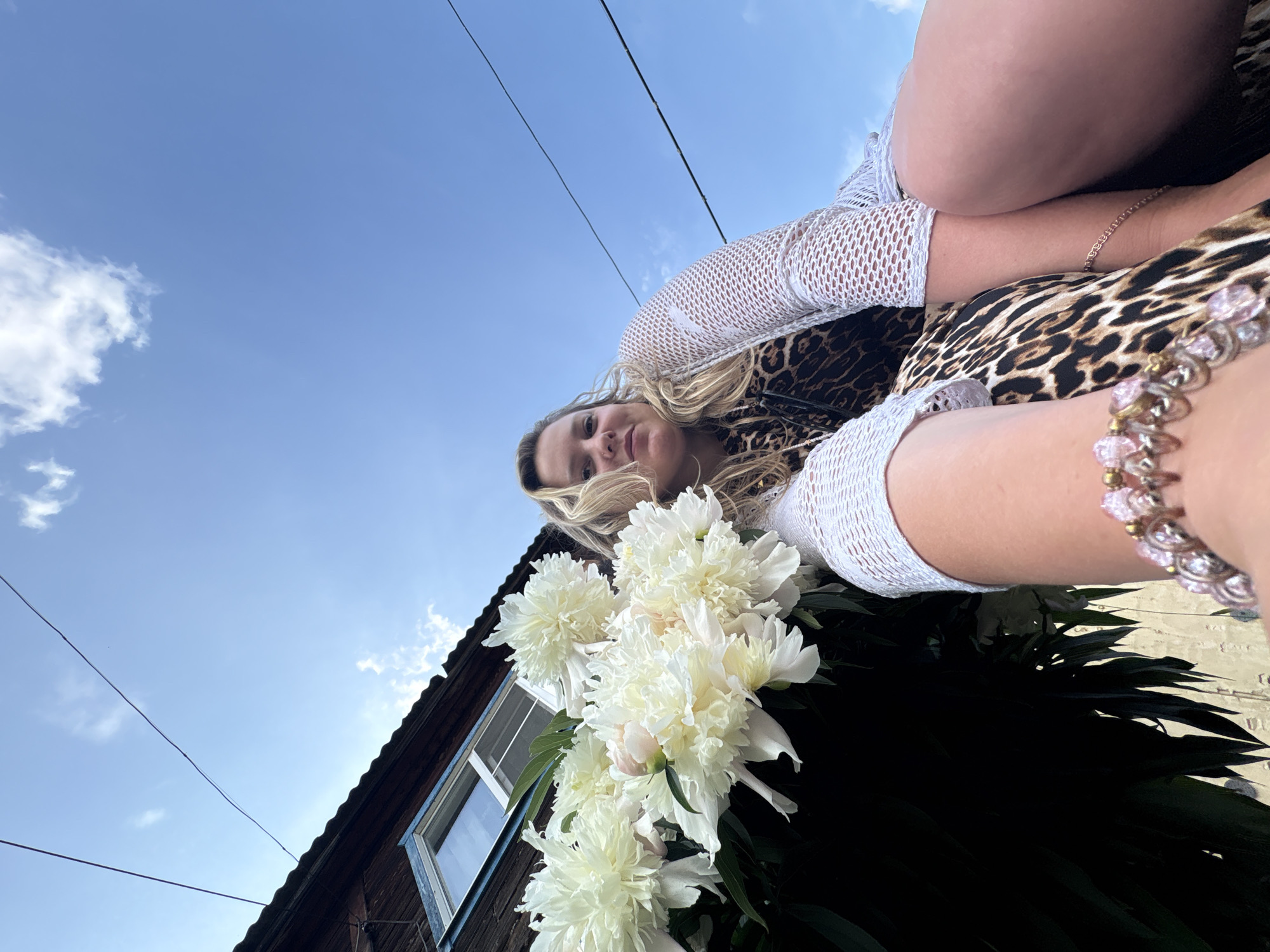 A low-angle, outdoor, medium shot captures a young blonde woman with fair skin, looking directly at the camera. She has light blonde, wavy hair and is wearing a white mesh top over a leopard-print dress. Her right arm is bent at the elbow, and she is wearing a bracelet with pink and silver charms. She appears to be sitting on something, as her legs are visible in the lower right of the frame, her right thigh and knee prominently displayed.
In the foreground, to the left, a large bouquet of white peonies is held by an unseen person, or possibly the subject herself, with dark green leaves. The bouquet partially obscures the lower left portion of the woman's face.
The background features a wooden building with a blue-trimmed window and a dark roof. Above the building, a clear blue sky is visible with a few wispy white clouds. Two thin black wires are stretched diagonally across the sky, suggesting the presence of electrical infrastructure, which is common in urban or semi-urban areas like Chaikovsky, Russia. The lighting indicates it is daytime, with the sun casting subtle shadows. The overall impression is that of a casual, outdoor portrait.