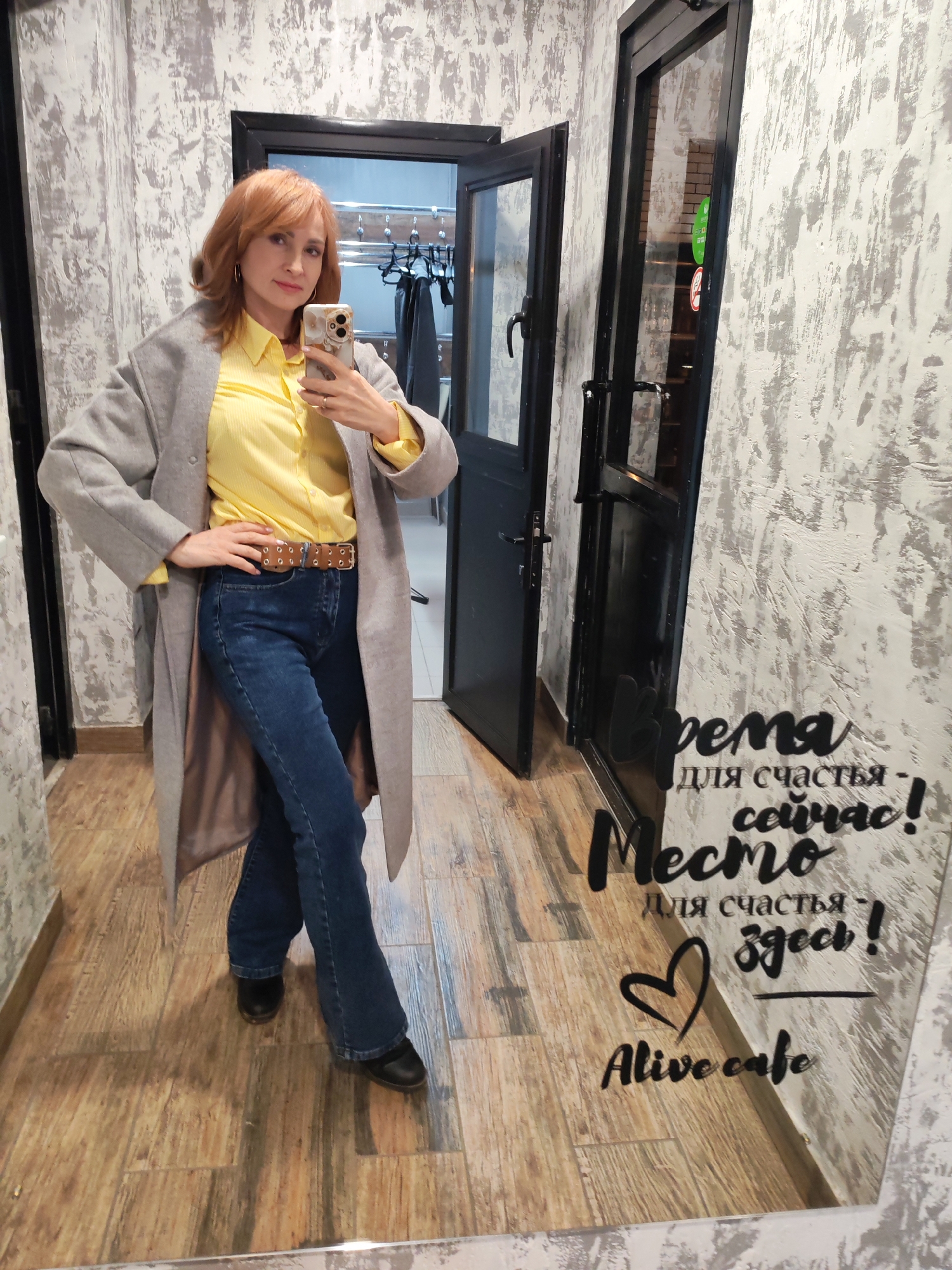 A woman takes a mirror selfie inside what appears to be a cafe or shop in Chaikovsky, Russia. She has reddish-brown, shoulder-length hair and is wearing a bright yellow collared shirt, dark blue jeans, a brown belt with silver eyelets, and a long, light grey coat. She holds a smartphone with a decorative case in her right hand, taking the photo, and her left hand is on her hip. She has a subtle smile and looks directly at her reflection/camera. Her shoes are black.
The background features a textured, light grey and white wall. To her right (viewer's left) is a dark-framed glass door, partially open, revealing a glimpse of an interior with a clothes rack. The floor is covered with light brown, wood-patterned tiles.
The large mirror on the right side of the image reflects the woman and the room. On the mirror, black lettering in Russian reads: "Время для счастья сейчас!" (Time for happiness now!) and "Место для счастья здесь!" (Place for happiness here!), followed by a heart symbol and "Alive cafe". The presence of these messages and the cafe name reinforces the commercial setting. The lighting is artificial, consistent with an indoor environment.