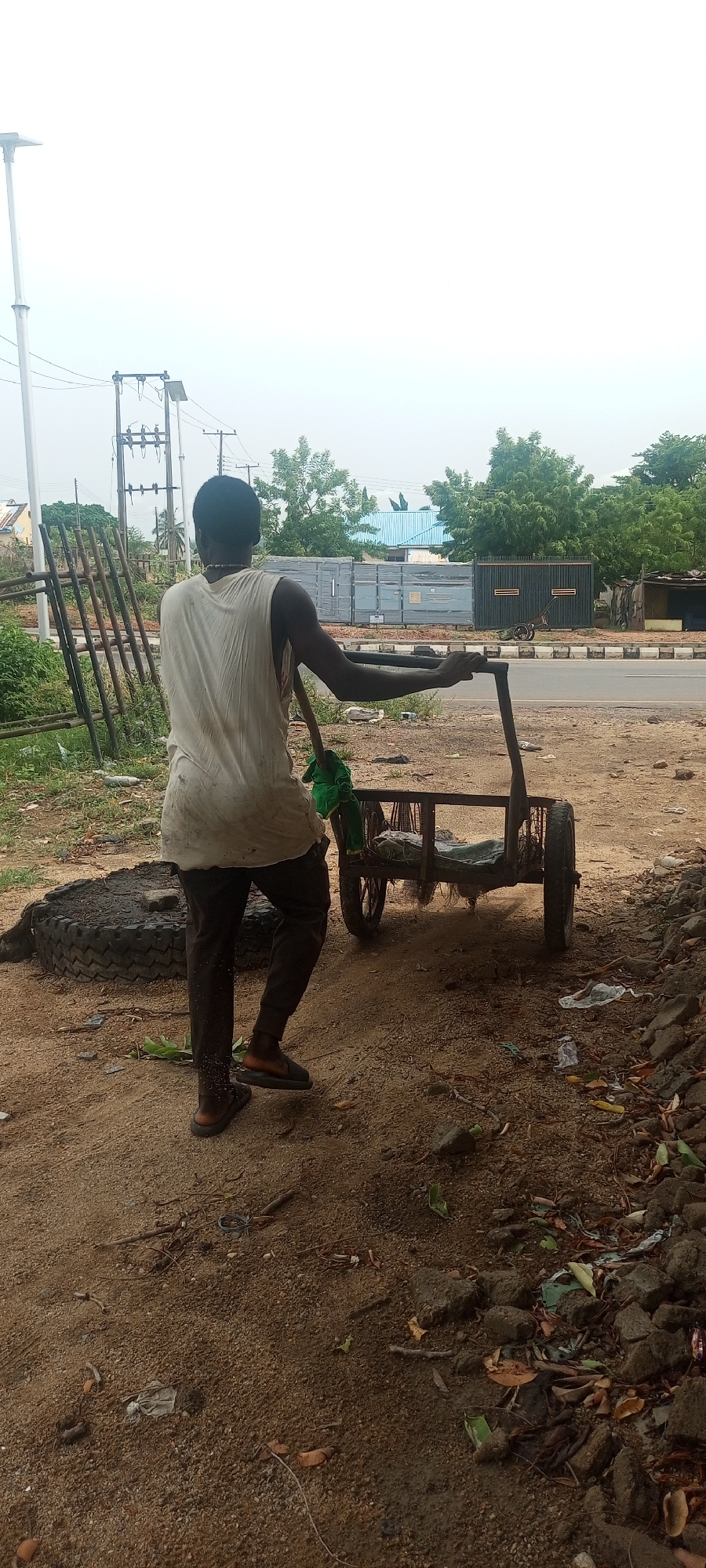 A man stands next to a four-wheeled cart on a sandy, trash-strewn ground in Zango-ndaji, Nigeria. The man, viewed from the back, wears a light-colored, sleeveless shirt and dark pants with sandals. He holds the cart's handle with both hands and a green object hangs from the cart's side. A large tire lies near a stack of rusty metal rods to the left of the man. The background features a road, trees, and buildings under an overcast sky. A utility pole is visible in the distance.