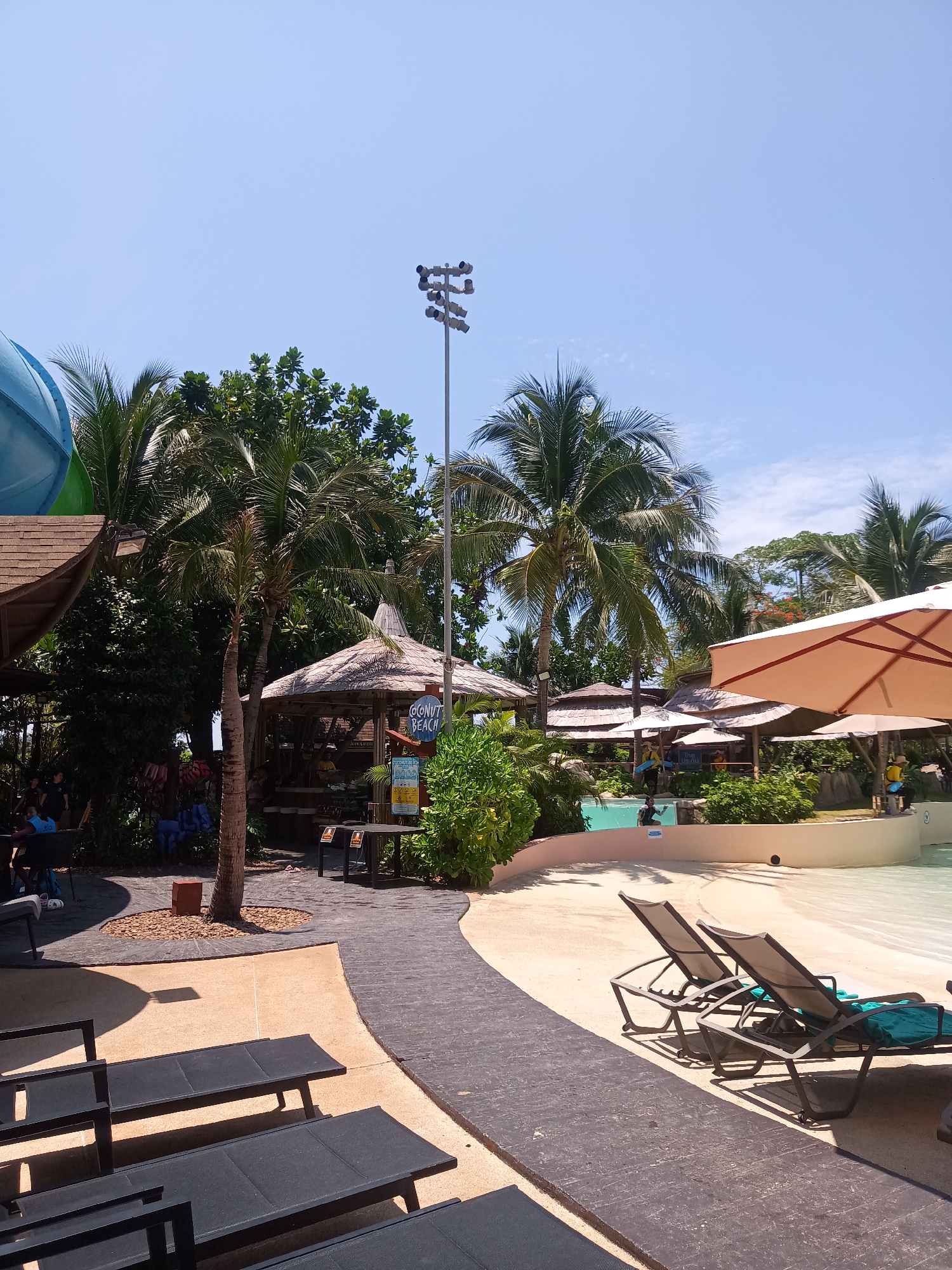 The image depicts a scene in Hua Hin City Municipality, Thailand, likely within a resort or water park. The location appears to be a recreational area with a pool and beach-like setting.
Foreground: Several dark-colored lounge chairs are arranged on a concrete surface. Two of them have teal-colored cushions. A black brick-paved walkway curves towards the right side of the image.
Midground: A shallow pool with simulated waves is visible. Lifeguards in uniform are present, indicating the presence of swimmers. There are several thatched-roof structures providing shade, along with large parasols. Lush tropical vegetation, including palm trees, is scattered throughout the area. One structure has a sign indicating "Coconut Beach."
Background: A tall light pole stands behind the trees. A portion of a blue water slide is visible on the left. The sky is clear and blue. People are visible under the shaded areas.