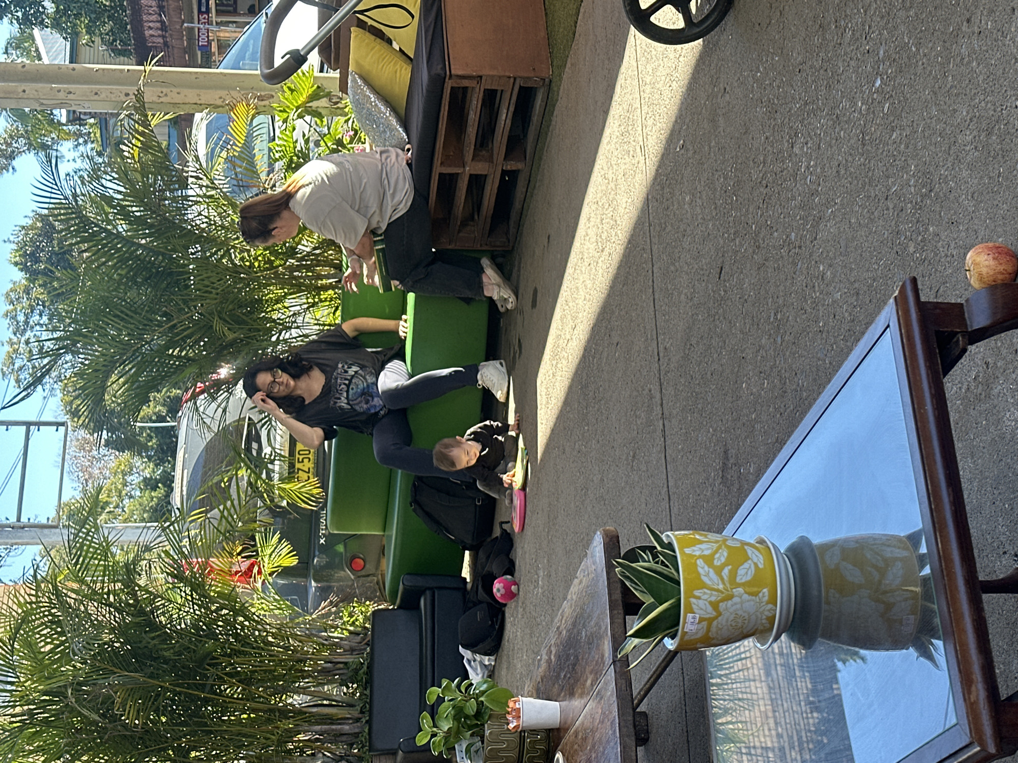 In Bellingen, Australia, two women are seated on an outdoor bench. One woman, with dark hair and glasses, wears a black t-shirt and dark pants, and is touching her hair. The other woman, with her hair in a ponytail, wears a light-colored t-shirt and dark pants and is gesturing with her hands. A baby is crawling on the ground between them, playing with a pink toy and a pink ball. The bench is made of green upholstered furniture and a wooden pallet base, with yellow and silver cushions. Behind the bench is a dark-colored SUV with a license plate ending in "50". To the left of the bench, a black couch is visible, along with potted plants and a dark backpack. In the foreground, a wooden table with a glass top holds a yellow potted plant. Sunlight casts shadows across the concrete ground. A stroller is partially visible on the right side of the image, and a red apple rests on the ground near the table. The environment suggests an outdoor seating area, possibly at a cafe or restaurant.