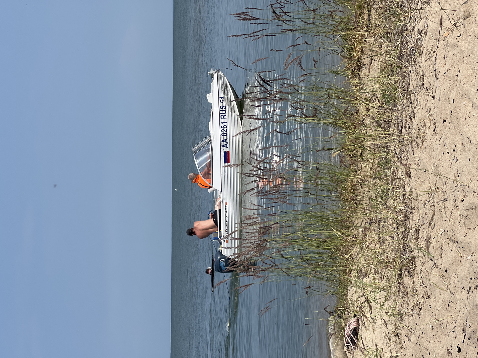 A white motorboat with the registration "AA 0261 RUS 54" is in the middle of the water, moving to the right. Two men are in the boat. One man, wearing an orange life vest, is in the passenger seat, and another man, shirtless, is in the driver's seat. A person is in the water behind the boat, appearing to be wakeboarding. In the foreground, tall grass and sand are visible, suggesting a beach or shoreline. The background shows a calm body of water under a clear blue sky. The overall scene depicts a recreational activity on the water.