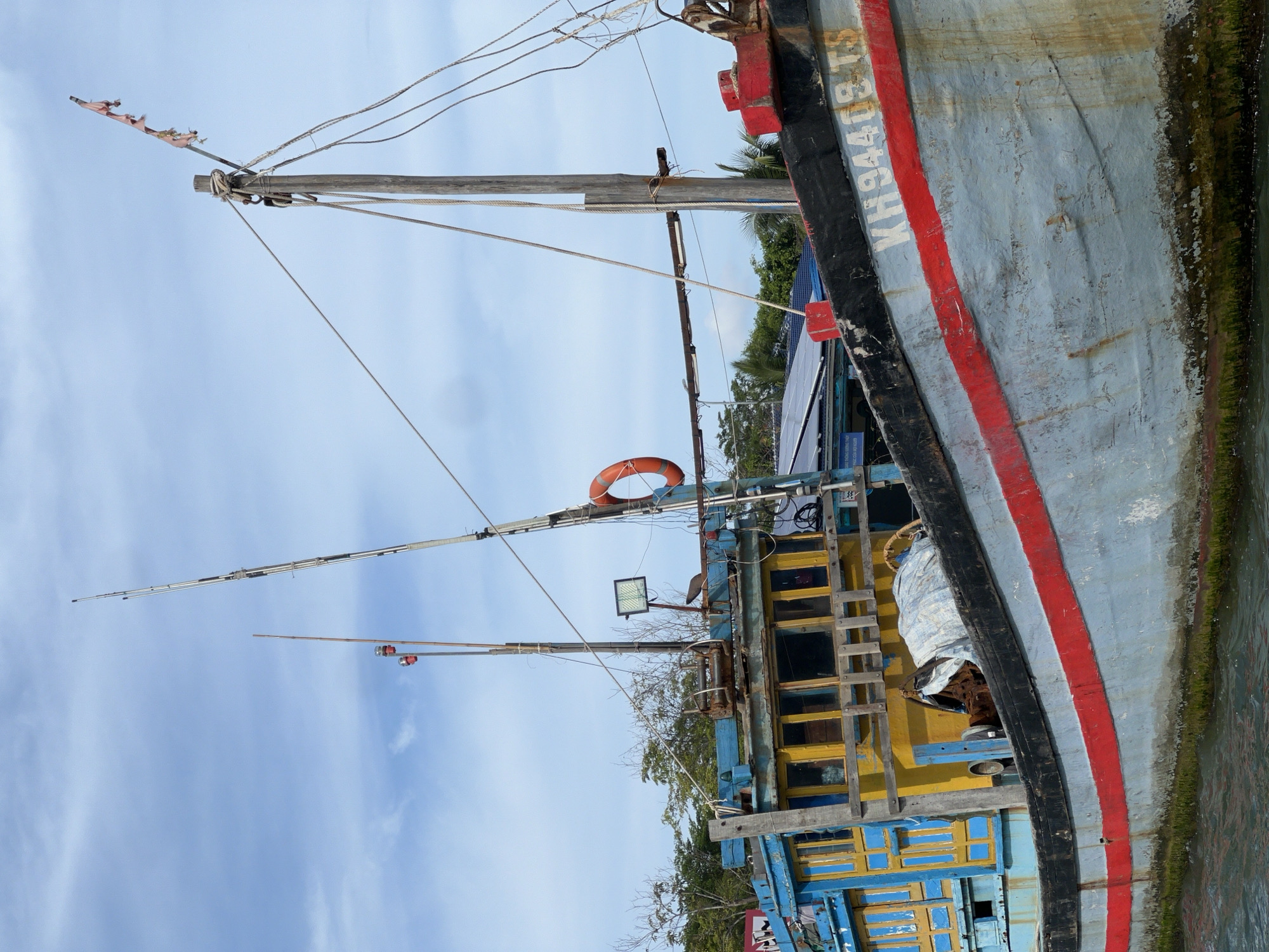 The image shows a close-up view of a boat in Nam Nha Trang Ward, Vietnam. The boat's hull is painted light blue with a red stripe and the registration number "KH94403TS" in white letters. The boat has multiple masts and rigging. A life preserver is visible. The structure of the boat includes blue and yellow painted sections, possibly cabins or storage areas. The boat is docked or in shallow water, with some green algae growth visible along the waterline. The sky is partly cloudy.