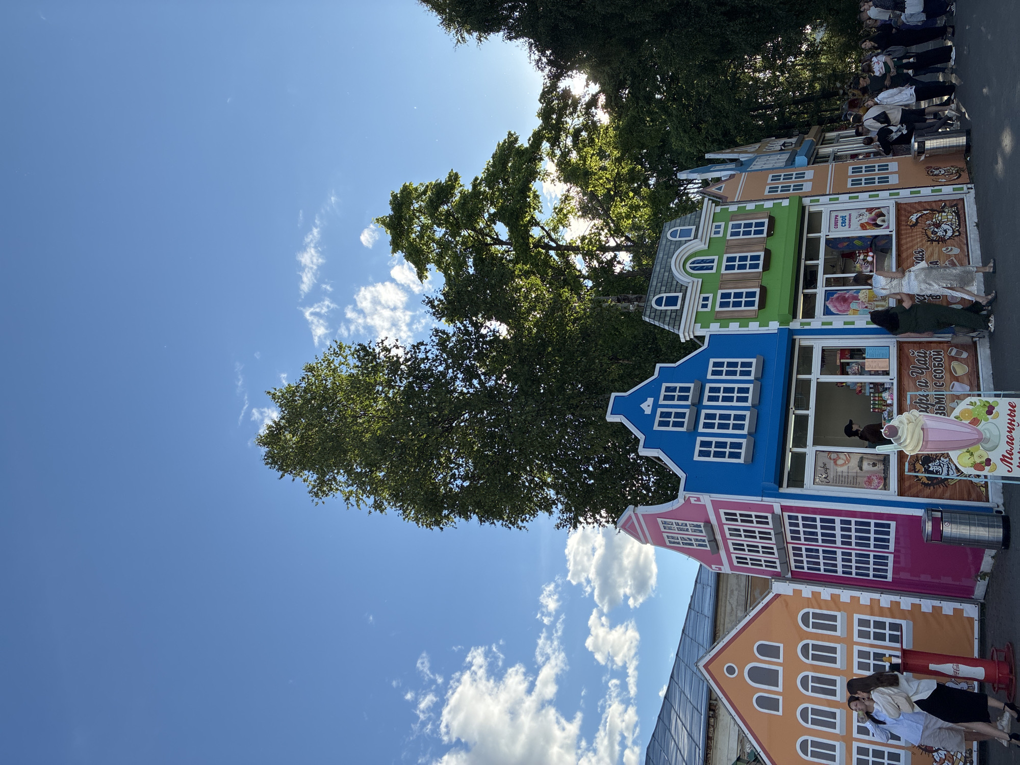 The image depicts a row of colorful, miniature buildings resembling Dutch houses, likely set up as stalls in an outdoor market or amusement park. The buildings are painted in bright hues of blue, green, pink, and orange, each with white-framed windows and decorative rooflines. The architecture mimics traditional Dutch style with pitched roofs and arched windows.
Several people are visible in the scene. In the foreground, two young women are embracing, appearing to share a tender moment. Further back, near one of the stalls, a woman in a silver dress stands, possibly interacting with someone inside the stall. A queue of people is also visible in the background, suggesting a popular attraction or service.
The stalls appear to be selling food or drinks. One stall has signage with images of ice cream and what looks like a milkshake. Another stall has text that appears to be in Cyrillic script, but it's not fully legible.
The setting is outdoors, with a clear blue sky and scattered white clouds. Lush green trees frame the top of the image, with sunlight filtering through the leaves, indicating it is daytime, likely late morning or afternoon. The overall mood seems cheerful and lively, suggesting a pleasant day for leisure activities. The image's orientation is sideways, with the buildings running horizontally across the frame.