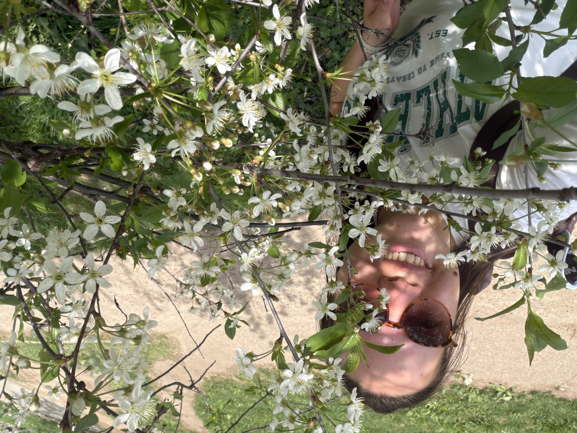 A woman with fair skin and dark hair, wearing sunglasses and a white t-shirt with a green graphic design, is positioned amongst branches of a blossoming tree with white flowers. The location appears to be outdoors, with a combination of grass and a paved surface in the background. Based on visual cues alone, the location cannot be definitively confirmed as Chaikovsky, Russia.