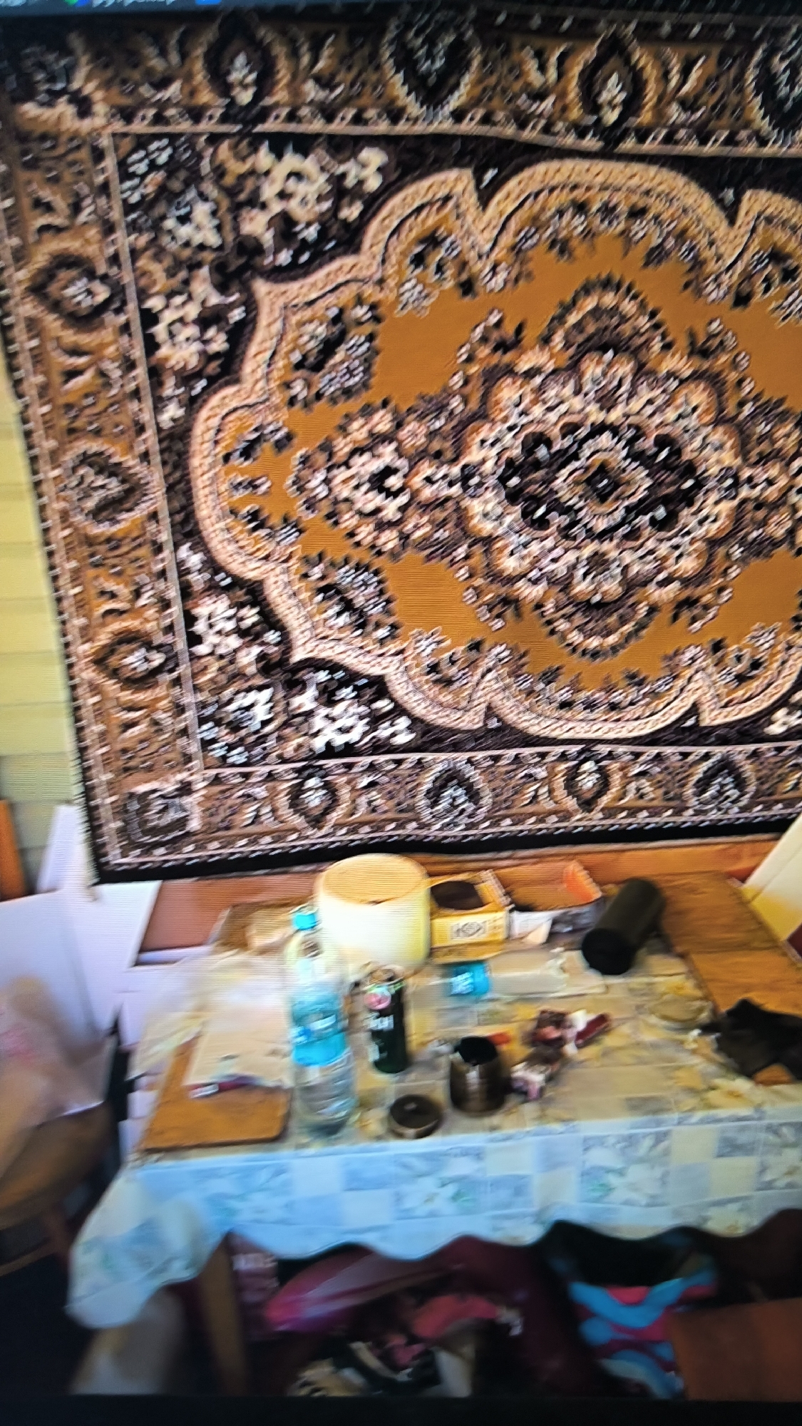 The image shows a dimly lit room, likely an apartment in Kovrov, Russia, with a large, ornate rug hanging on the wall as a tapestry. The rug is predominantly gold and brown with intricate floral patterns. In the foreground, a table is set with a checkered tablecloth. On the table, several objects are visible, including a plastic bottle of water, a can of soda, a small metal container, a wooden cutting board, and what appears to be a box. The lighting is poor, and the overall scene suggests a lived-in space. There are no people visible in the image. The presence of the rug suggests a potentially older or traditional decor style.