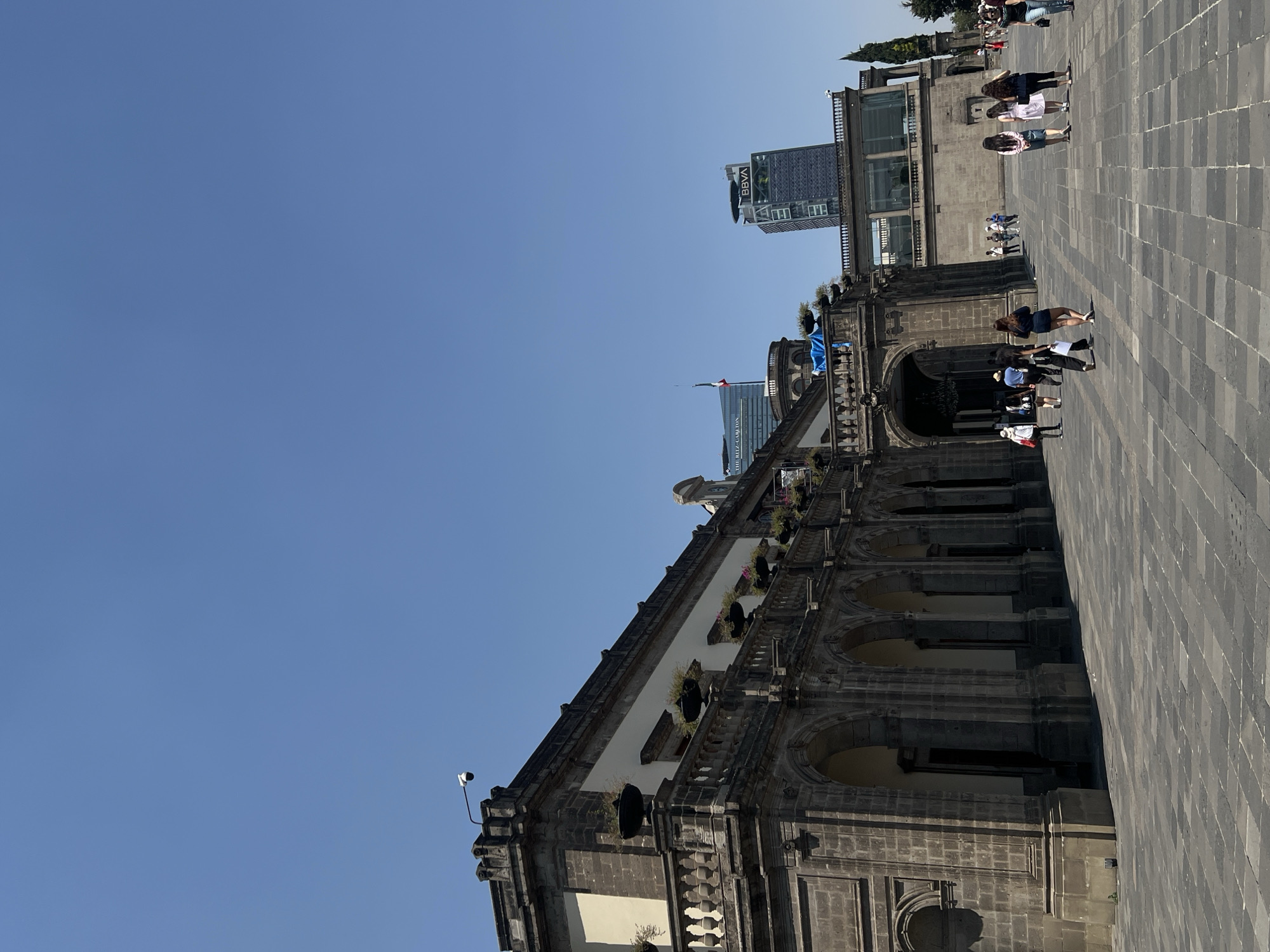 An outdoor scene captures a large, bustling public square in Mexico City under a clear, bright blue sky. The image is taken from a low angle, looking up and across the square.
In the foreground and dominating the left side is a grand, historic stone building, possibly a cathedral or a palace, characterized by its dark grey stonework, arched windows, and balconies adorned with planters and greenery. A modern security camera is mounted on its roofline. The square itself is paved with dark grey rectangular stones, creating a grid pattern.
Numerous people are seen walking and standing across the square. They appear to be casually dressed and are engaged in everyday activities, either moving through the plaza or observing their surroundings. Their presence suggests a busy, typical day.
In the background, beyond the historic structure, several modern skyscrapers rise into the sky. One prominent high-rise clearly displays the "BBVA" logo near its top, while another, partially visible, bears the text "THE RITZ-CARLTON." A flag, likely the Mexican flag, is seen flying from a pole adjacent to one of these modern buildings, further indicating the location.
The time of day appears to be sunny, likely midday or early afternoon, with clear weather casting shadows on the paved ground and structures. The overall atmosphere is one of a vibrant urban space where historic architecture meets contemporary development.