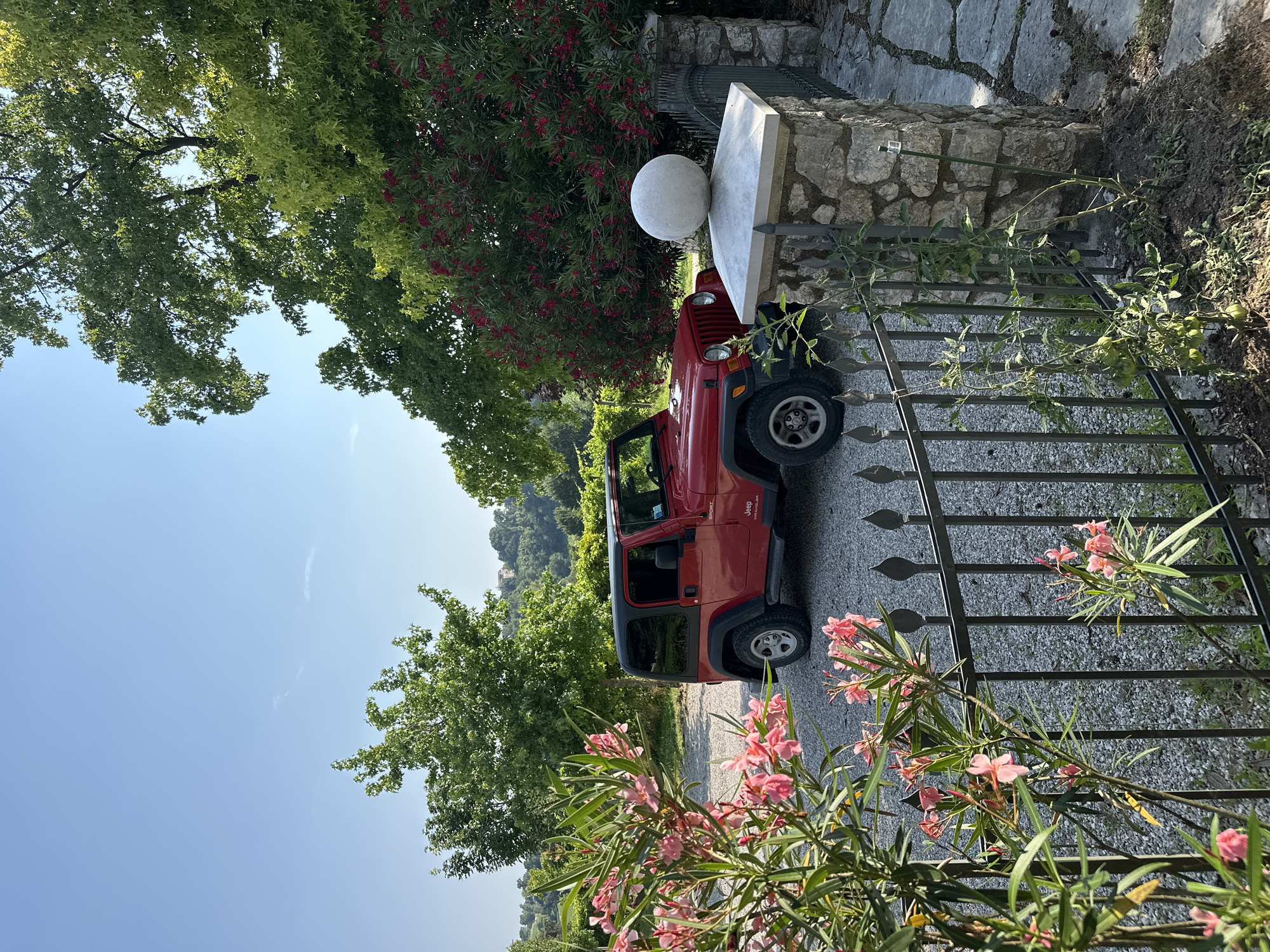 The image depicts a red Jeep Wrangler parked on a gravel driveway. The Jeep is positioned near a stone pillar with a spherical sculpture and a flat stone slab. Behind the pillar and Jeep, there are lush green trees and bushes with red flowers. In the foreground, pink oleander flowers bloom, obscuring a black metal fence. The scene appears to be outdoors with a clear blue sky. The time of day is likely daytime, given the bright sunlight casting shadows. The presence of oleander flowers and the style of stonework suggest a Mediterranean or Italian setting. There is no activity taking place; it is a still scene. No people are visible in the image. The word "Jeep" is visible on the side of the vehicle, along with "SPORT" and what appears to be "WRANGLER".