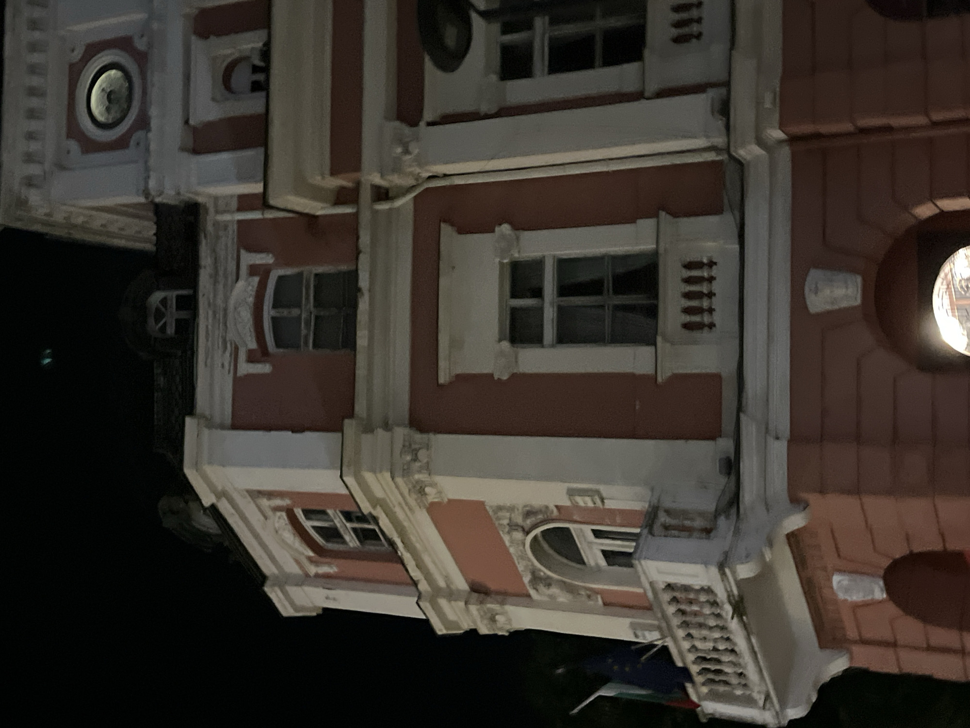 The image shows an exterior view of a building at night. The building appears to be made of brick and has ornate architectural details, including decorative moldings, columns, and arched windows. Several windows are visible, some with multiple panes. The building's facade is painted a reddish-brown color with white trim. In the foreground, there is a dark area that could be a street or a sidewalk, and a flag with green and white stripes is partially visible. The overall scene suggests an urban or historic setting. The time of day is clearly night, as indicated by the darkness and the artificial lighting within some of the windows. There are no people visible in the image, and no specific activity or event is taking place. The image is oriented vertically, with the bottom of the building appearing at the top of the frame.