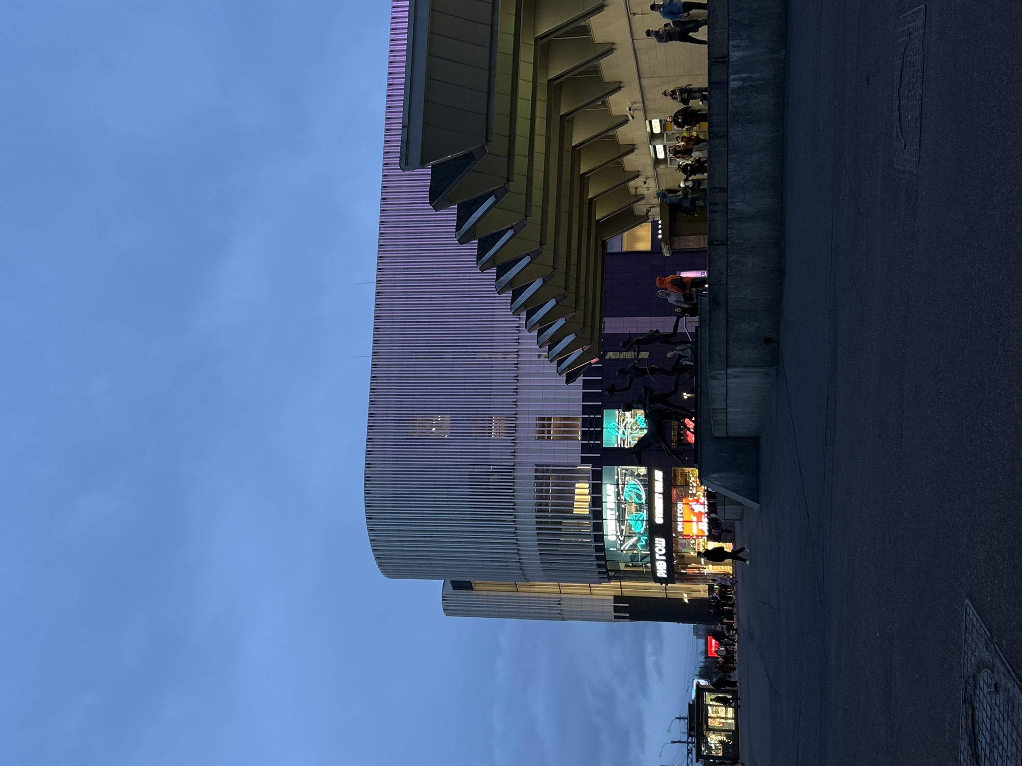 This image captures a modern urban scene in Saint Petersburg, Russia, during twilight or early evening. The sky is a clear, light blue, suggesting the transition from day to night.
The main subject is a contemporary multi-story building on the left, featuring a distinctive facade. Its upper section is characterized by numerous vertical, light-colored slats or fins, which appear to reflect a subtle purple light in some areas. Below this, the building showcases several large, brightly lit digital or neon signs. One prominent sign displays the mirrored text "RIV GOSH" and "Street wear" in English, referencing a well-known Russian cosmetics retailer and clothing. Adjacent to these illuminated signs, a dark, abstract sculptural figure or statue is visible. Higher up on the building, there are angular, stepped structures, possibly part of the building's upper levels or awnings.
To the right, a dark asphalt street or wide pedestrian area stretches out, featuring a rectangular manhole cover. A low concrete wall separates this ground level from an elevated platform or terrace of the building.
Numerous people populate the scene, engaged in typical urban activities. On the elevated platform, several individuals are walking or standing, their forms largely silhouetted against the background light. At street level, pedestrians are seen walking past the brightly illuminated storefronts and signs, with one person in a distinct orange hoodie visible near the dark sculpture. The artificial lights of the building and its advertisements glow vibrantly, contrasting with the fading natural light. The overall atmosphere is that of a calm yet active urban environment in the evening.