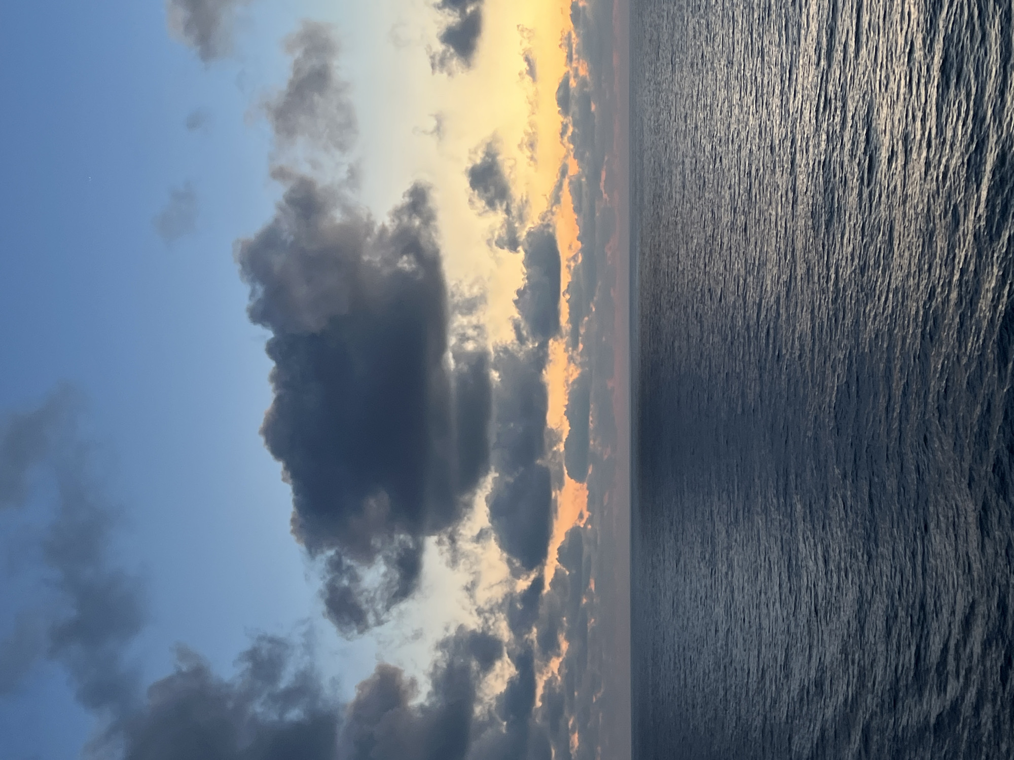 The image depicts a vast expanse of dark blue ocean under a twilight sky. The horizon line is clearly defined, separating the water from the sky. The sky is a gradient of colors, transitioning from a pale blue at the top to warmer hues of orange and yellow near the horizon. Dark, heavy clouds are scattered across the sky, some casting shadows on the water below. A single, faint star is visible in the upper right quadrant of the sky. The surface of the ocean is rippled, indicating a gentle movement of water, with subtle reflections of the sky's colors visible on its surface. The scene is serene and appears to be captured from a boat or elevated vantage point overlooking the sea. There are no people, structures, or any indication of a city visible in the image.