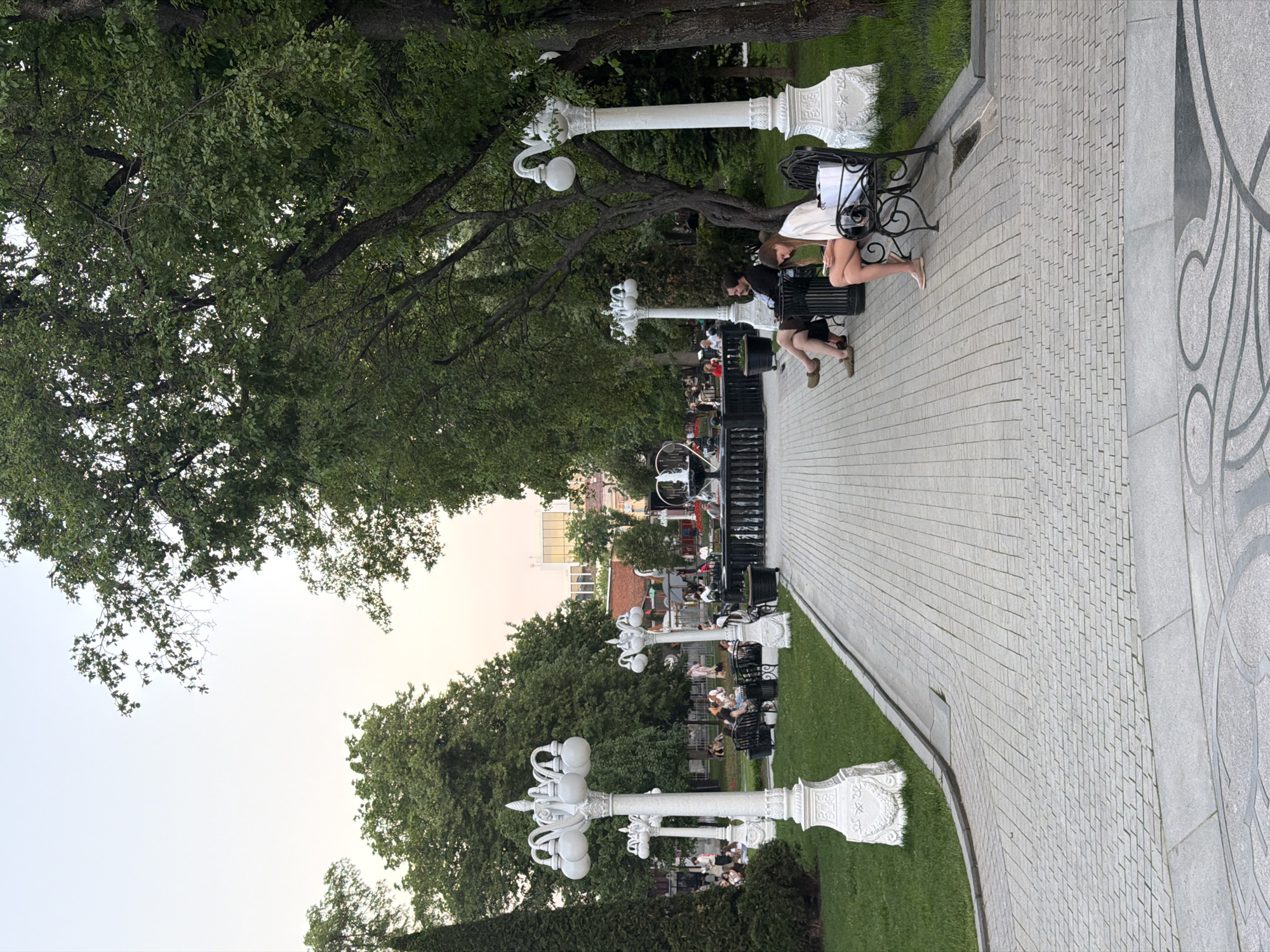A street scene captured in Moscow, Russia, features a paved walkway bordered by green grass and trees. Along the walkway, there are white, decorative street lamps and black benches where people are seated. Some individuals are seen interacting with each other on the benches. The weather appears clear, with daylight illuminating the scene.