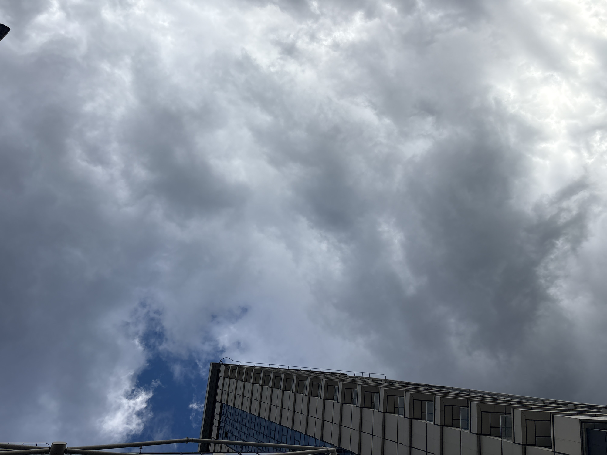 The image presents a striking low-angle perspective of a modern building against a dramatic, mostly cloudy sky. The building, possibly a high-rise located in Sofia, Bulgaria, occupies the lower right quadrant, showcasing a contemporary architectural style. Its facade is characterized by a repetitive, geometric pattern of deep-set windows or balconies, rendered in light-colored material with dark frames, creating a staggered, sculptural effect. Some sections of the building feature reflective dark glass, mirroring the grey tones of the sky.
The sky above is largely dominated by extensive, dark grey clouds, giving an overcast and somewhat moody impression. However, a significant patch of clear blue sky, interspersed with brighter white clouds, is visible towards the lower left, suggesting a break in the cloud cover or partially clearing conditions. The overall lighting is diffused, typical of a cloudy daytime scene. In the extreme lower left, a metallic structure composed of horizontal pipes or beams is partially visible, possibly scaffolding or part of a railing. No people are visible, nor are there any specific activities taking place. There is no discernible text in the image. The scene captures an urban environment under a dynamic and atmospheric sky.