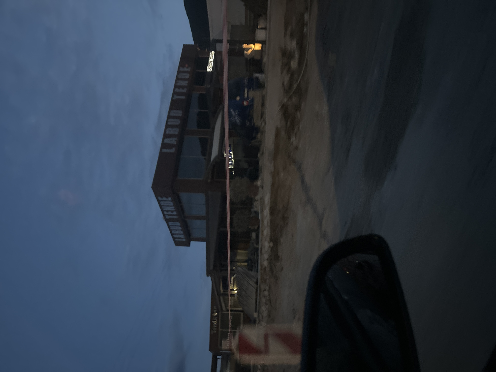 The image shows a building with the sign "LABUD TENDE" in Lastva Grbaljska, Montenegro. The photo was taken from a vehicle; a car mirror is partially visible on the right side. The sky is overcast. The road appears wet. Construction material is visible near the building.