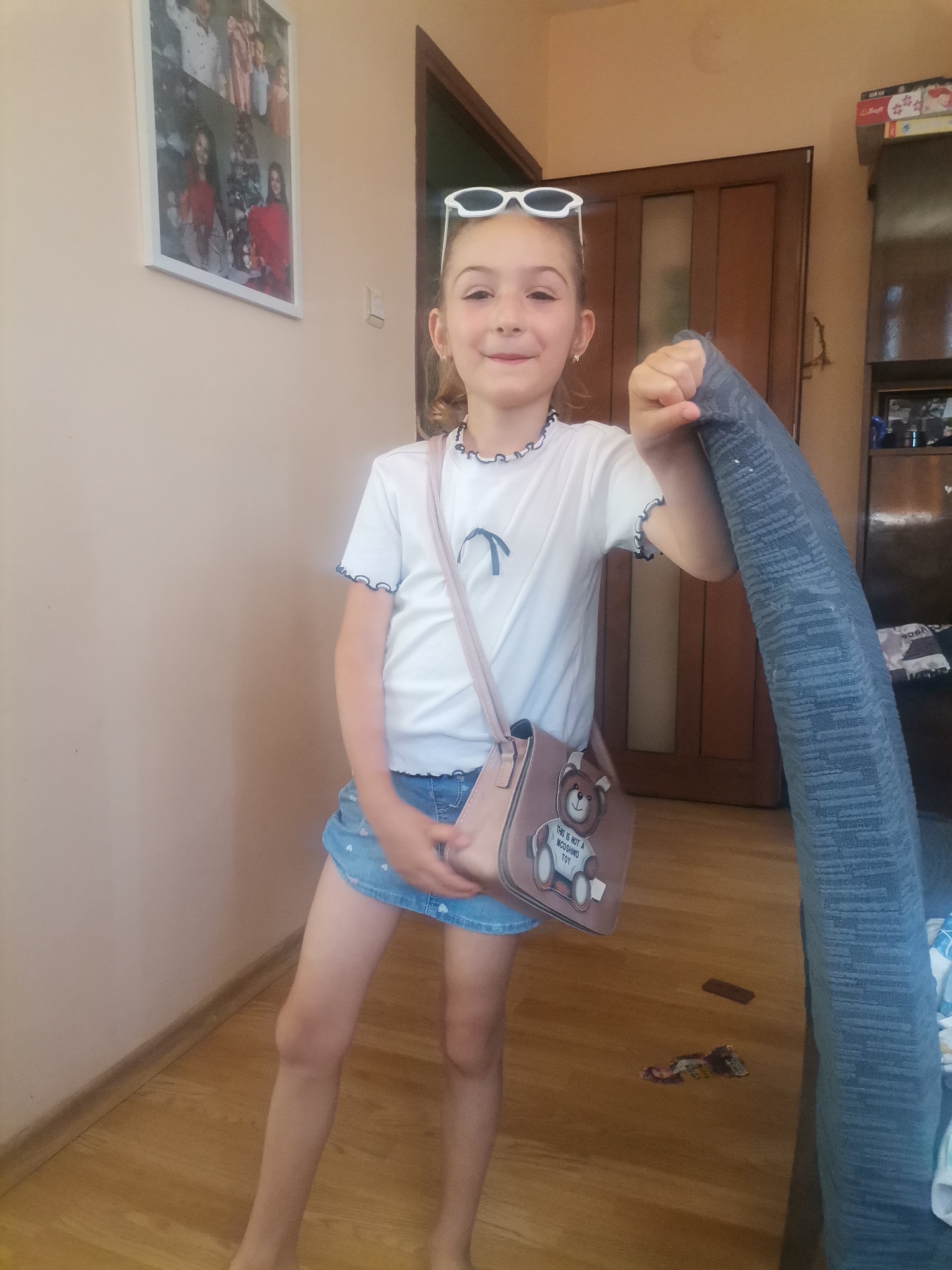 A young girl, appearing to be around 6-8 years old, stands in an indoor setting, possibly a home in Obrochishte, Bulgaria. She is wearing a white t-shirt with blue trim, a denim skirt with a subtle heart pattern, and sunglasses perched on her head. A light pink crossbody bag with a teddy bear motif is slung over her shoulder. The girl is holding a rolled-up blue textured item in her right hand, which could be a towel or blanket. She is smiling at the camera, conveying a playful and happy emotion. The background shows a light-colored wall with a framed collage of family photos, a wooden door, and a closet with visible items. The lighting suggests it is daytime. On the wooden floor, there are a few small objects, including what appears to be a wrapper and a dark rectangular item. The bag has text that reads "THIS IS NOT A MOSCHINO TOY".