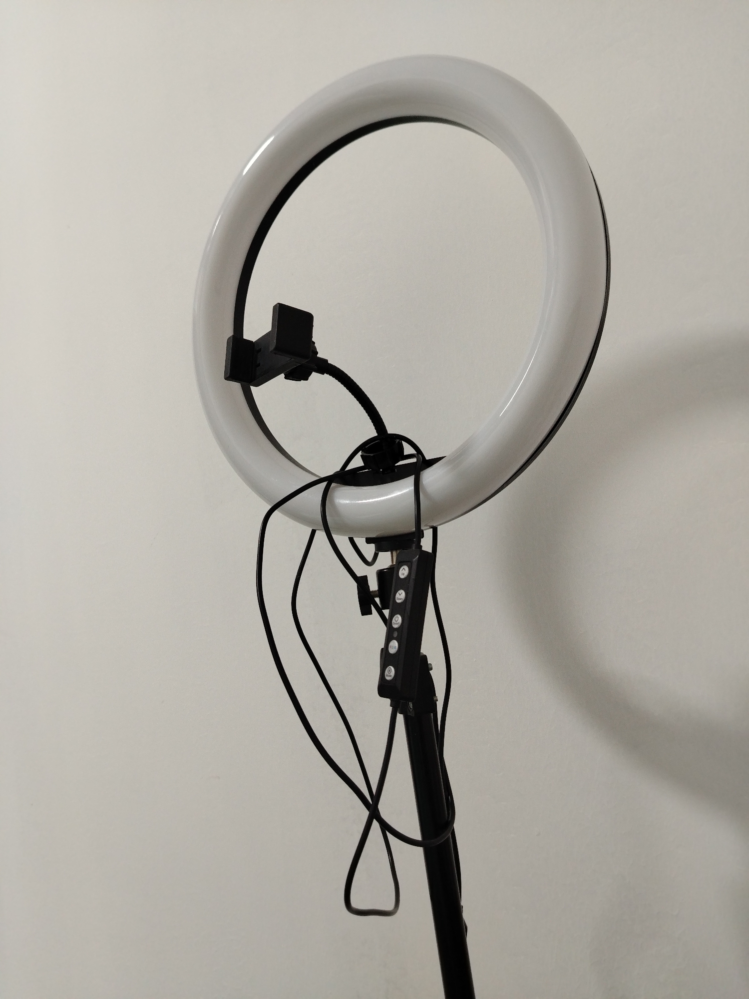 The image shows a ring light on a stand against a plain, off-white wall. The ring light is white and circular, with a black interior edge. A black phone holder is attached to the ring. A black control panel is visible on the stand, along with a collection of black cables. The stand is black and extends vertically. The location is not discernible from the image. There are no people visible.
