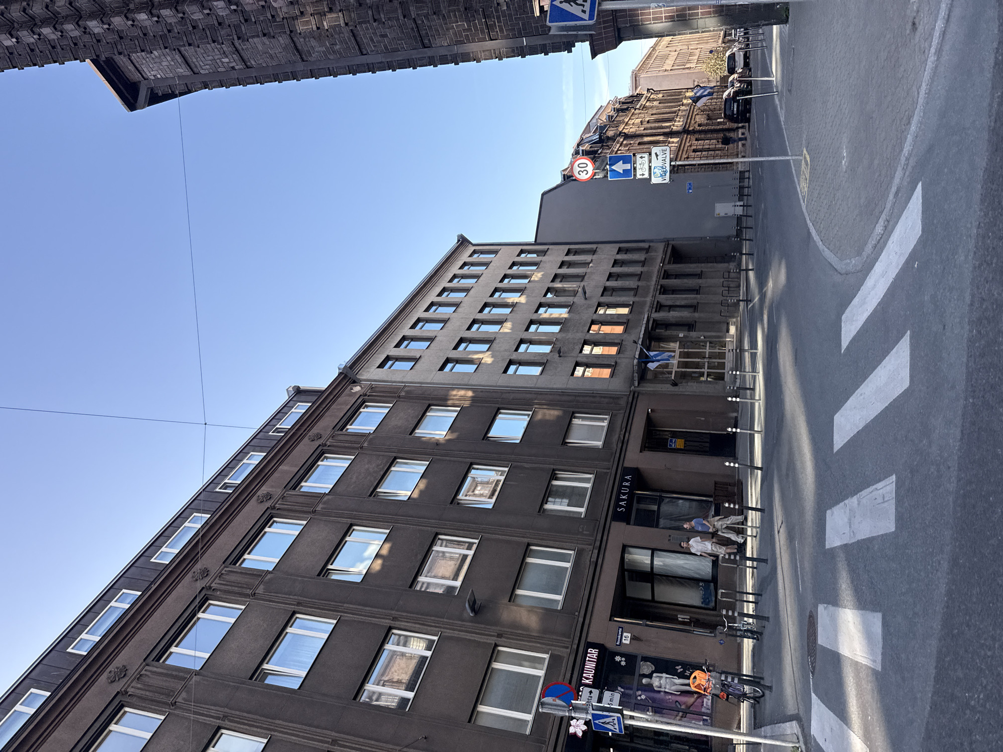 This is a street scene in Tallinn, Estonia, likely from the city center, featuring several multi-story buildings and a pedestrian crossing. The building on the left is a dark-colored, four-story structure with numerous windows and decorative elements. The building to its right is a lighter gray, also four stories, with a repeating pattern of windows. Further down the street, more buildings with classical architectural features are visible.
Several traffic signs are present. A blue and red circular sign with a diagonal red bar indicates a prohibition. A white square sign with a blue border depicts a pedestrian crossing symbol. Another circular sign shows a red border and a white background with "30" indicating a speed limit. Below this, a sign displays an upward arrow, a bicycle symbol, and a pedestrian symbol, suggesting a route for these modes of transport. A sign with a camera and text "VIDEOVALVE" is also visible.
On the sidewalk in front of the dark building, there are two bicycles parked. Two men are standing near the entrance to a shop labeled "SAKURA". In the foreground, a pedestrian crossing is clearly marked with white stripes on the asphalt road. The sky is clear and blue, suggesting daytime.