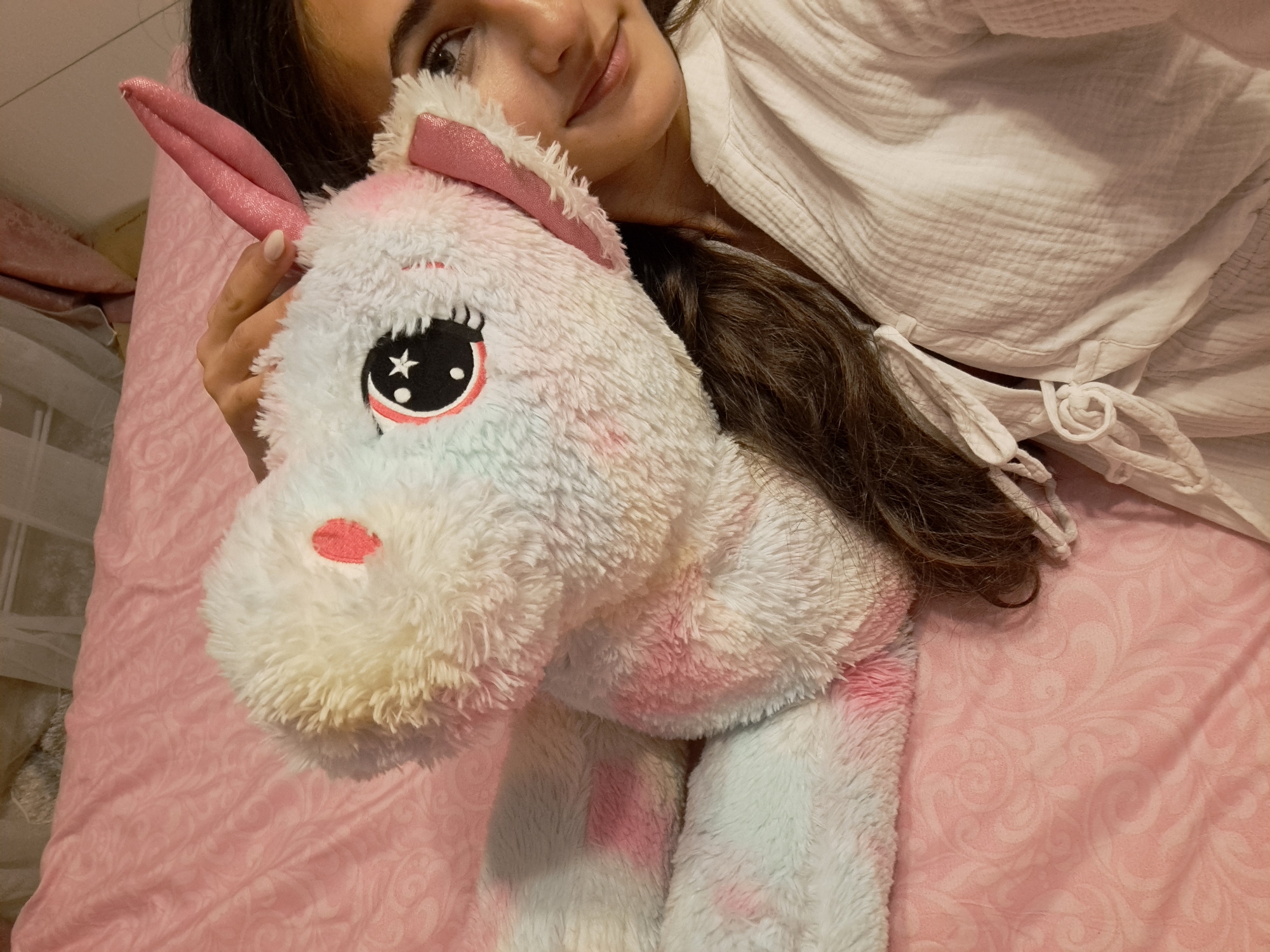 A person is depicted from an overhead perspective, positioned on a pink patterned bedspread in the city of Unknown, Unknown. The individual, whose right side of the face is visible, has dark hair, fair skin, and a subtle smile. They are wearing a white, textured, long-sleeved garment with tie closures. Their right hand, adorned with light-colored nail polish, rests on a large, plush, white unicorn stuffed animal with pink and light blue color accents. The unicorn features prominent black eyes with a white star and crescent moon design, pink inner ears, a pink horn, and a pink cheek marking. The unicorn is positioned close to the person's face, partially obscuring the left side of their head. The background consists primarily of the pink bedspread with a subtle relief pattern.